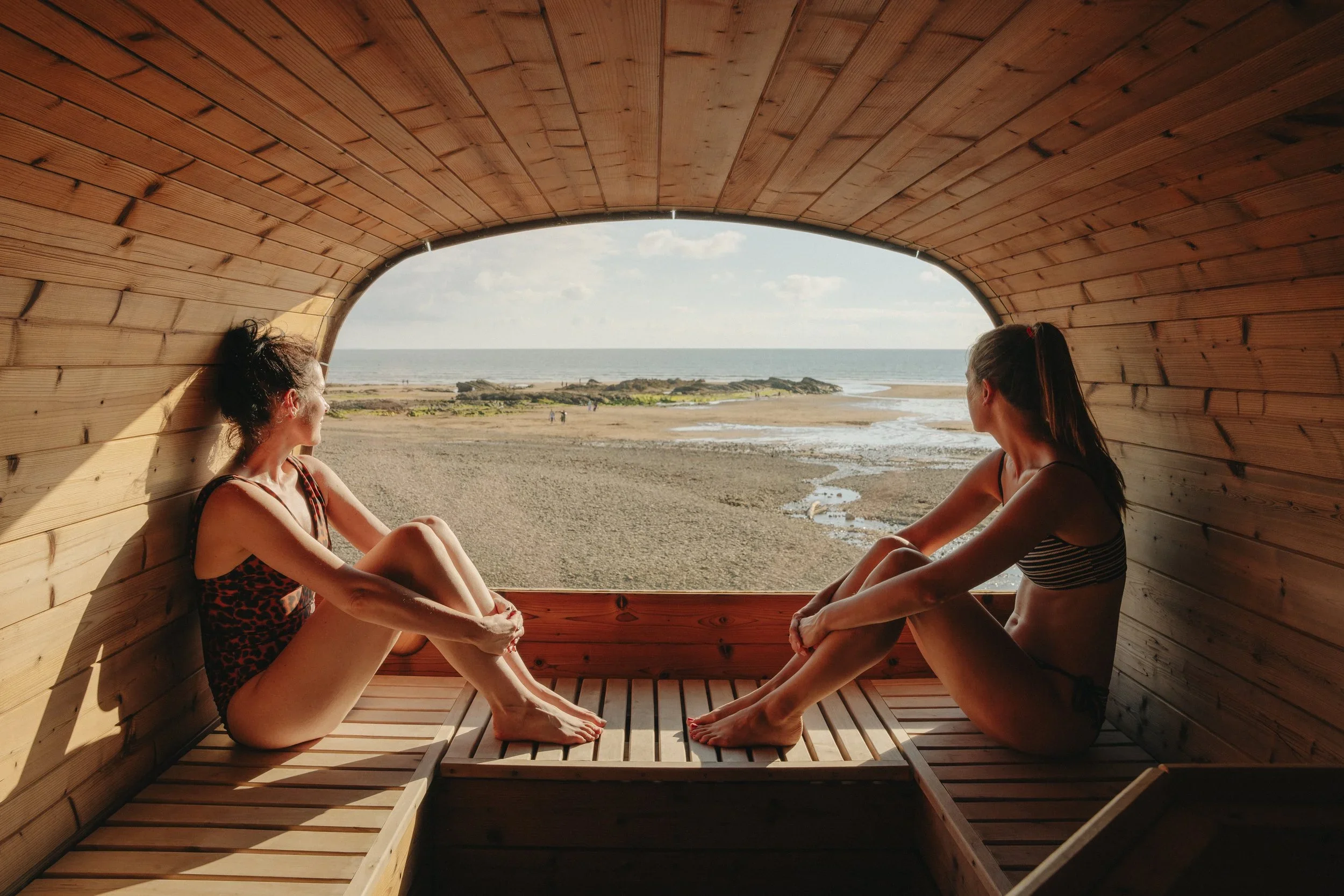 Two women sitting cross-legged inside a wooden, tunnel-like structure overlooking a beach and ocean.