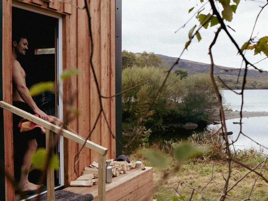 A man standing at the entrance of a wooden cabin by a lake, with trees and hills in the background, partially obscured by branches and leaves in the foreground.