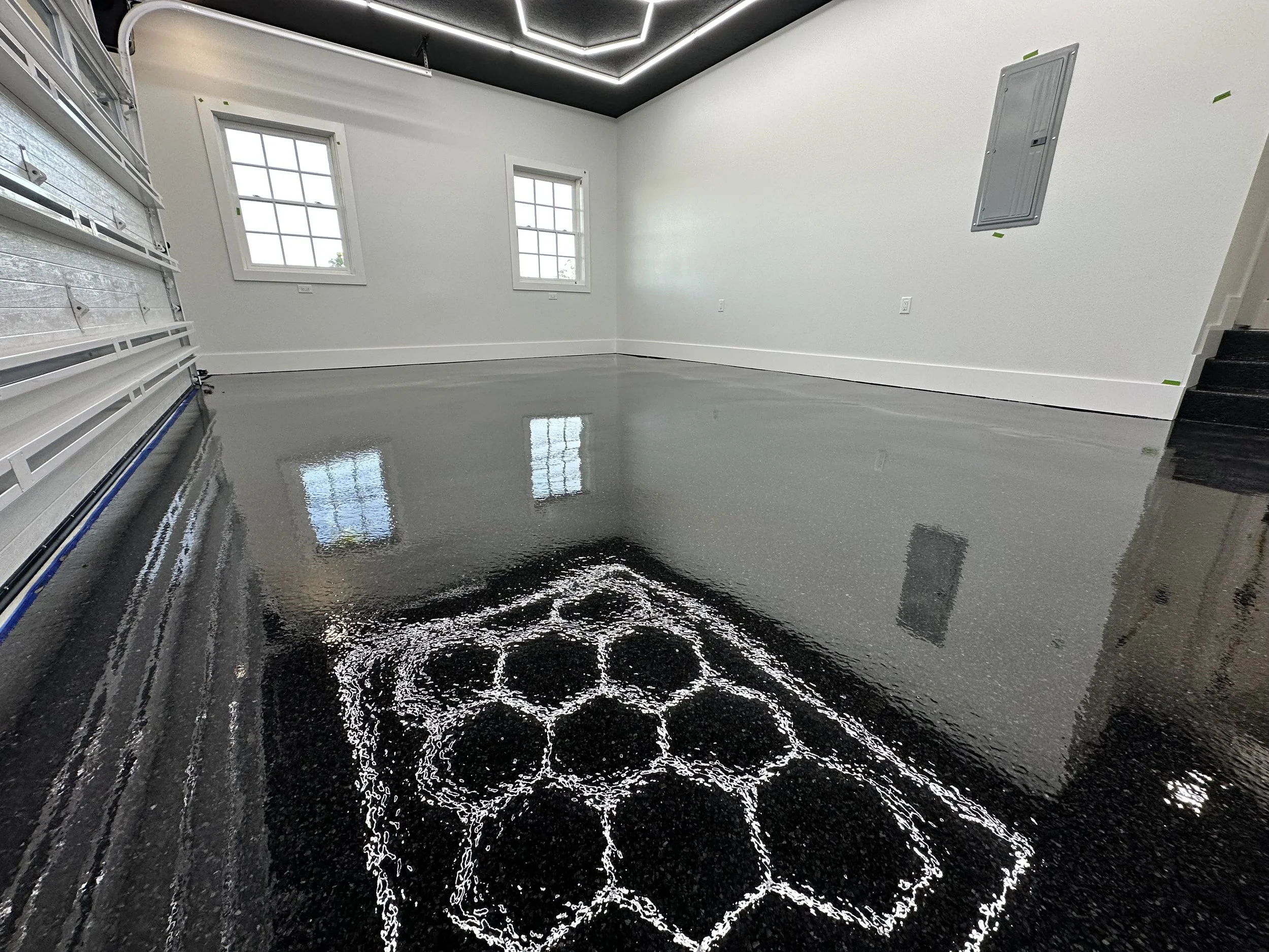 Empty garage with a freshly poured, glossy dark gray epoxy floor coating, reflecting the white walls, windows, and ceiling lights.