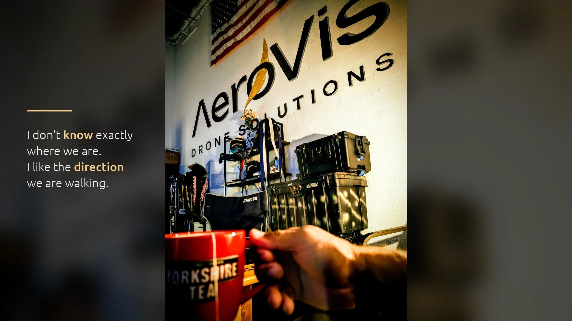 Kevin Padilla holds a coffee mug inside the AeroVis Drone Solutions shop in Sunrise, Florida, with the AeroVis wall mark, an American flag, and stacked equipment cases behind him.