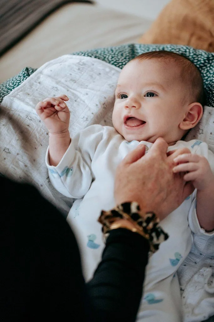 Un bébé souriant, allongé au sol avec sa maman qui interragit avec lui.