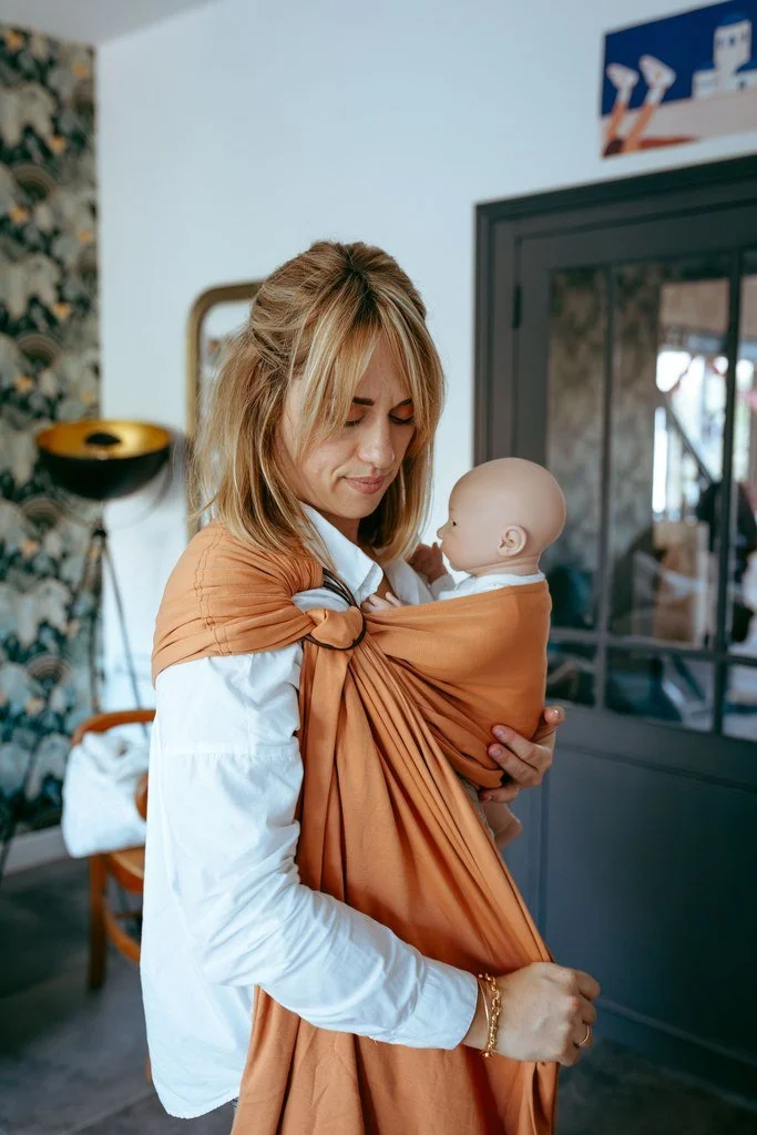 Une femme tenant un bébé dans un porte-bébé orange, dans une pièce intérieure.