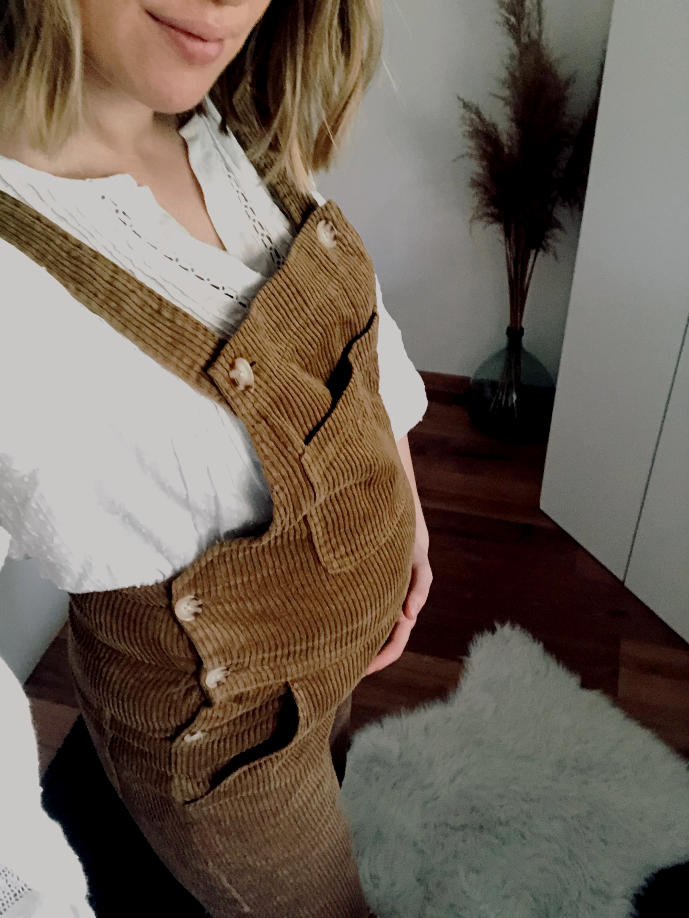 Femme avec un ventre rond, portant une chemise blanche et un pull en velours côtelé marron clair avec boutons, dans un intérieur avec une plante en vase et un meuble blanc.