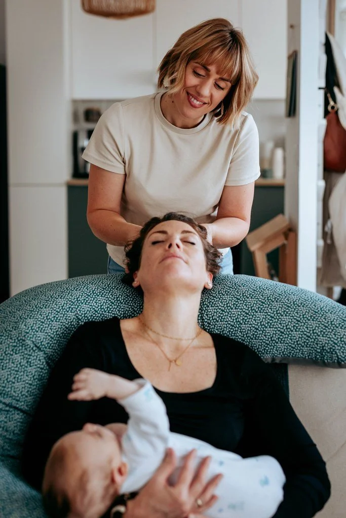 Une femme souriante en train de donner un massage de tête à une autre femme allongée sur un canapé, tenant un bébé dans ses bras.