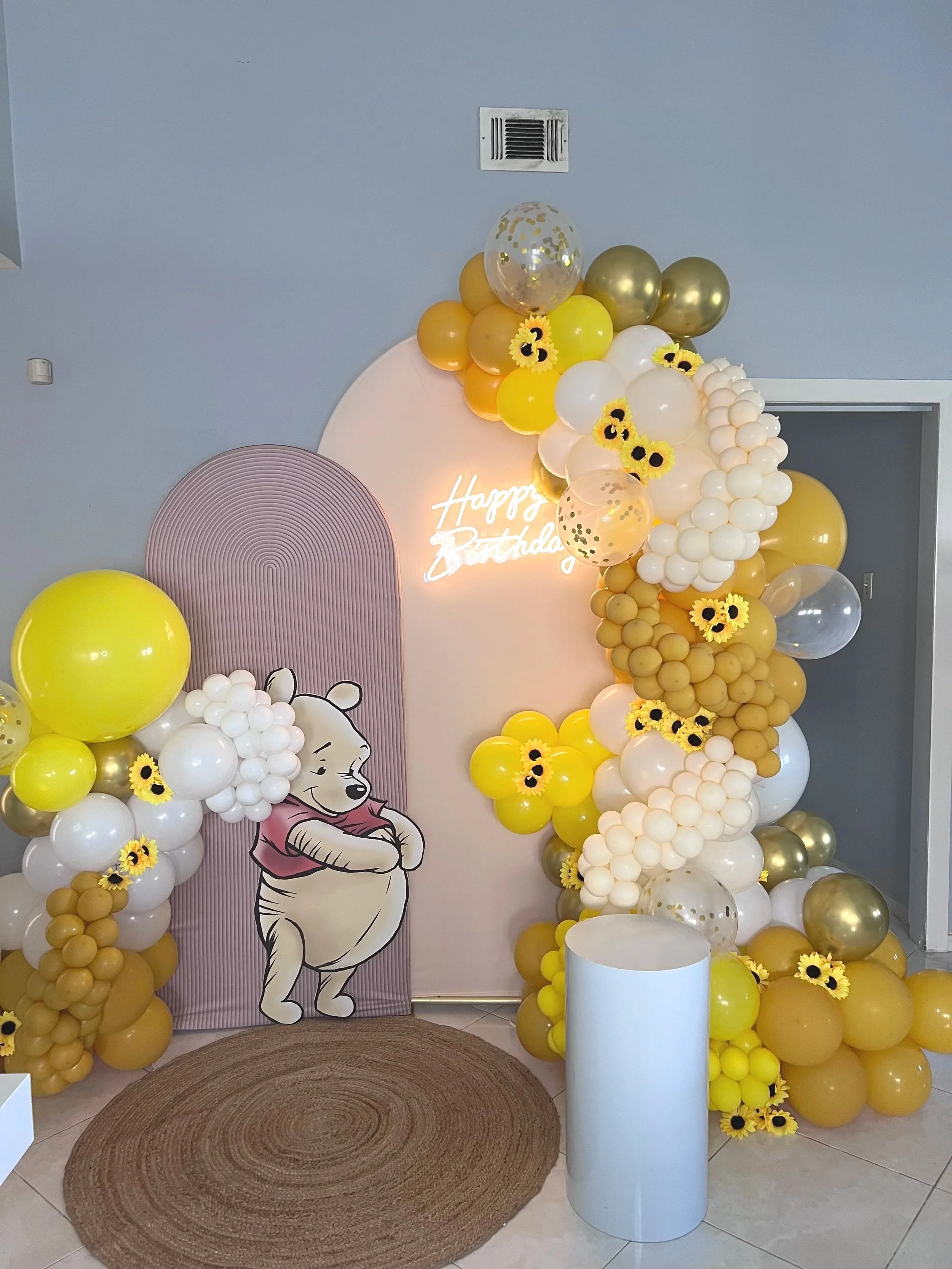 Balloon arch with white, yellow, gold, and transparent balloons and sunflower accents, surrounding a tan backdrop with a Winnie the Pooh figure and a neon sign that says 'Happy Birthday'.