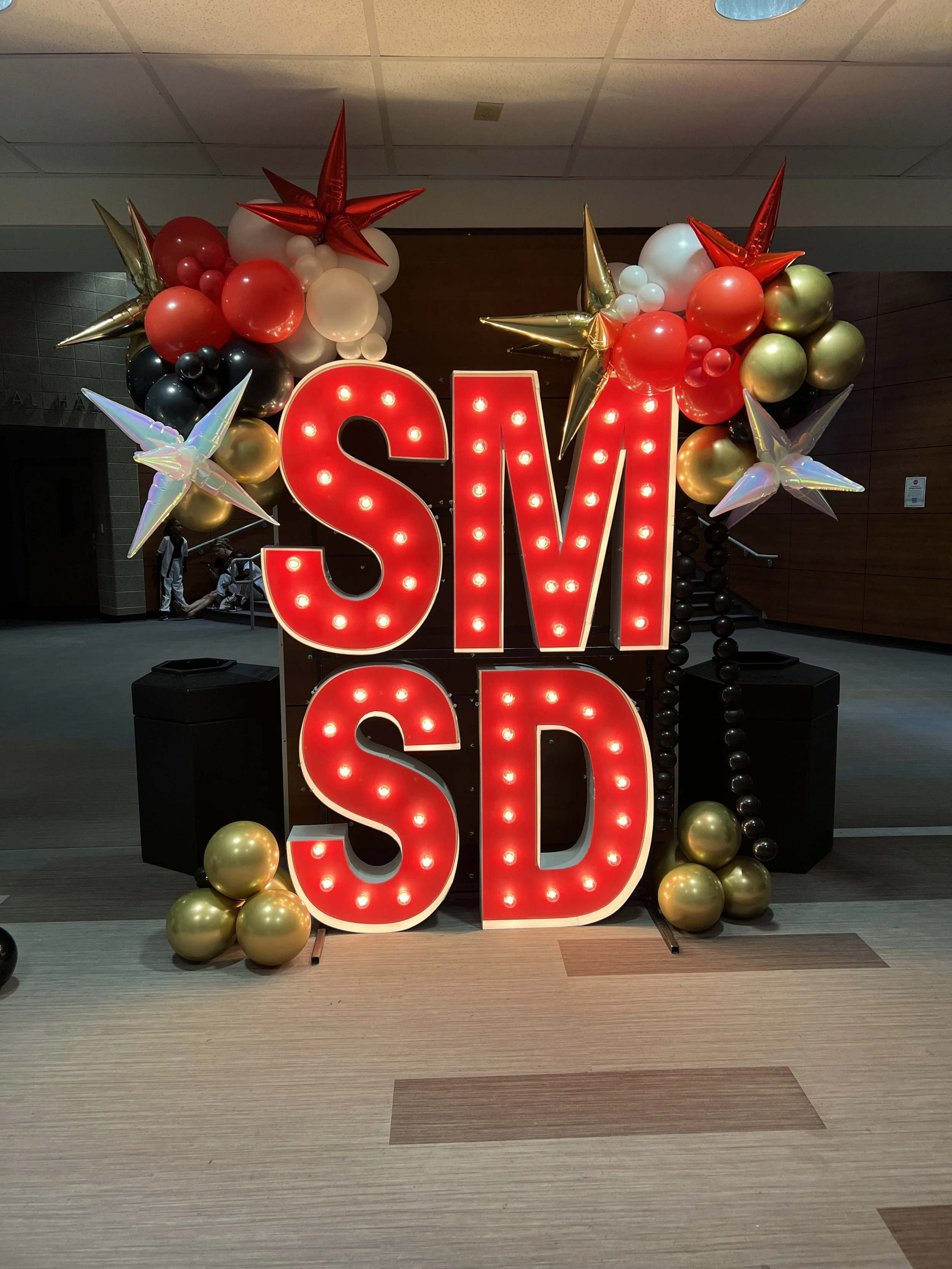 Shirley McPhail School of Dance marquee letters "S", "M", "S", "D", with red, gold, and white balloon garland with red, gold and iridescent starburst balloon accents.