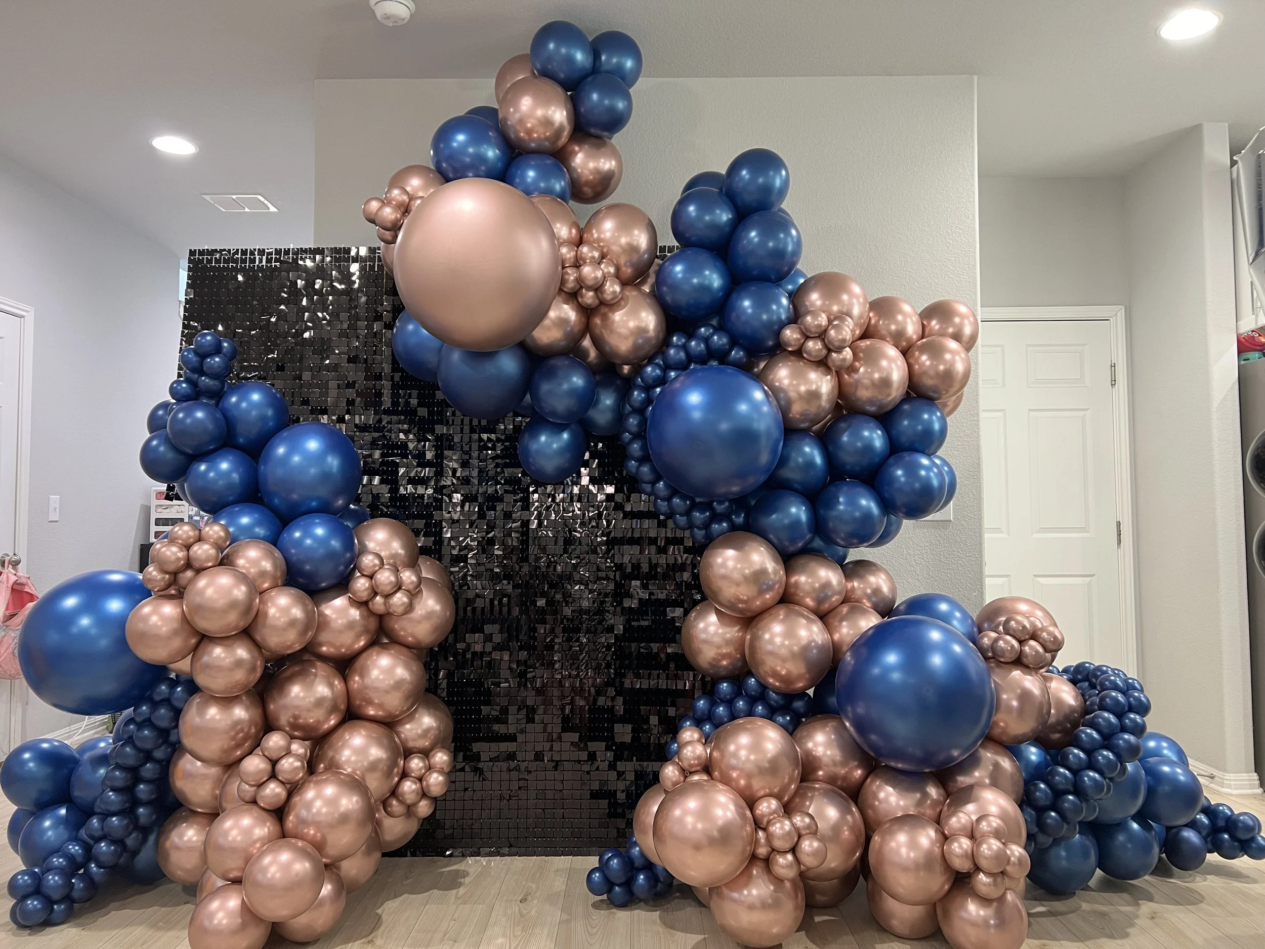 A decorative balloon arrangement with large metallic pink and blue balloons clustered around a black mirrored backdrop.