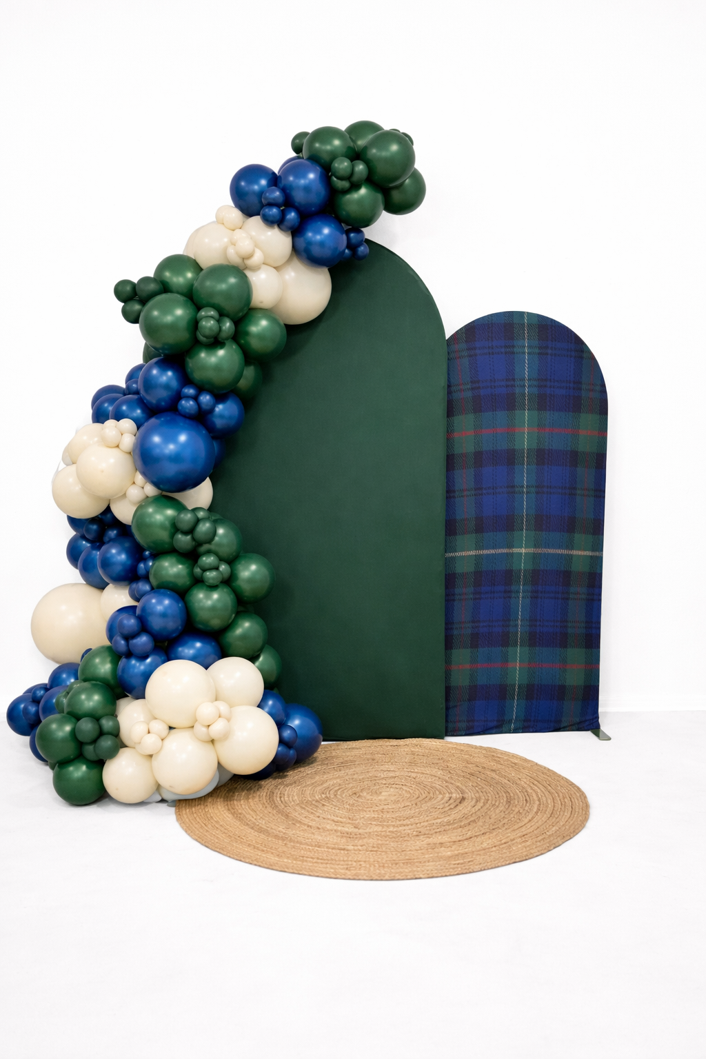 Decorative display with a dark green and plaid backdrop, green, blue, and cream balloons, and a round jute rug.