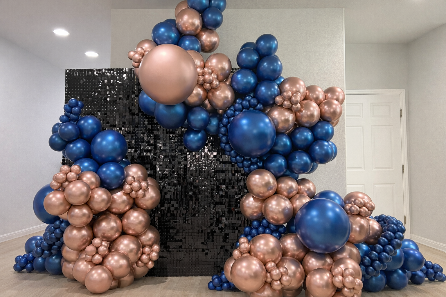 Decorative balloon arrangement with blue and metallic pink balloons against a black reflective backdrop in a room with gray walls .