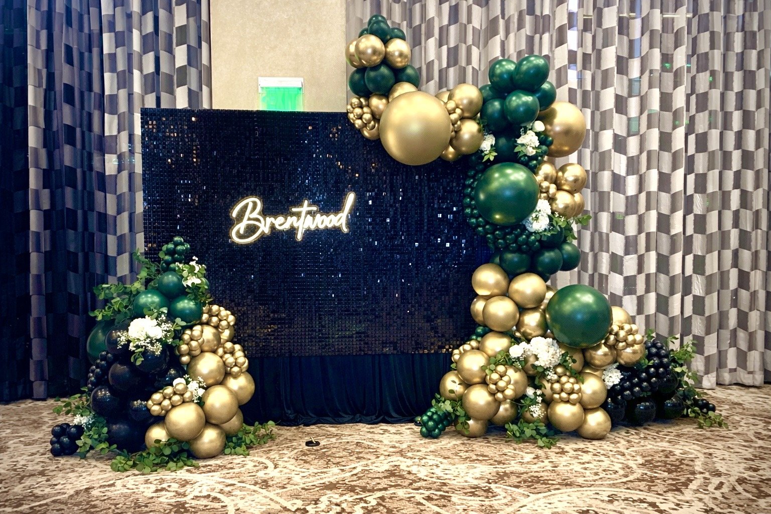 Gold, black, and Green ballon Garland on a black shimmer wall that has a Brentwood neon sign.