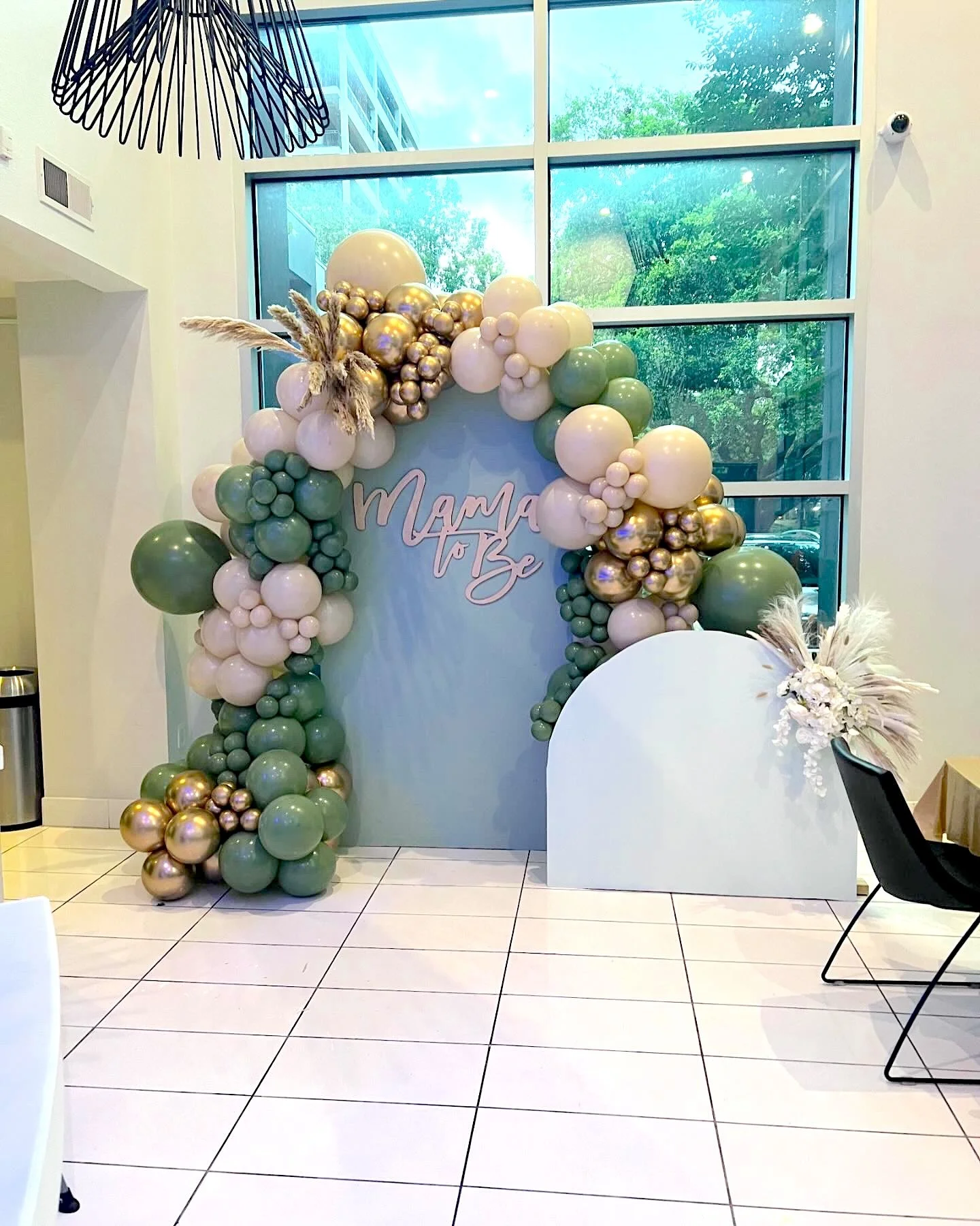 Balloon decoration arch with green, white, and gold balloons, with a sign that says "Mama To Be" in the center, inside a modern room with large windows.