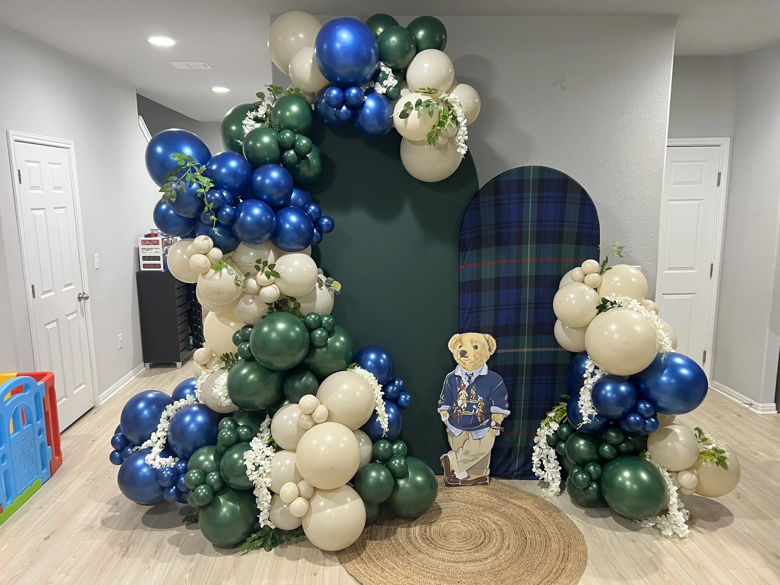 Decorative balloon arrangement in blue, green, and cream colors, forming an arch and columns around a plaid backdrop with a teddy bear cutout, in a room with white walls and light wood flooring.