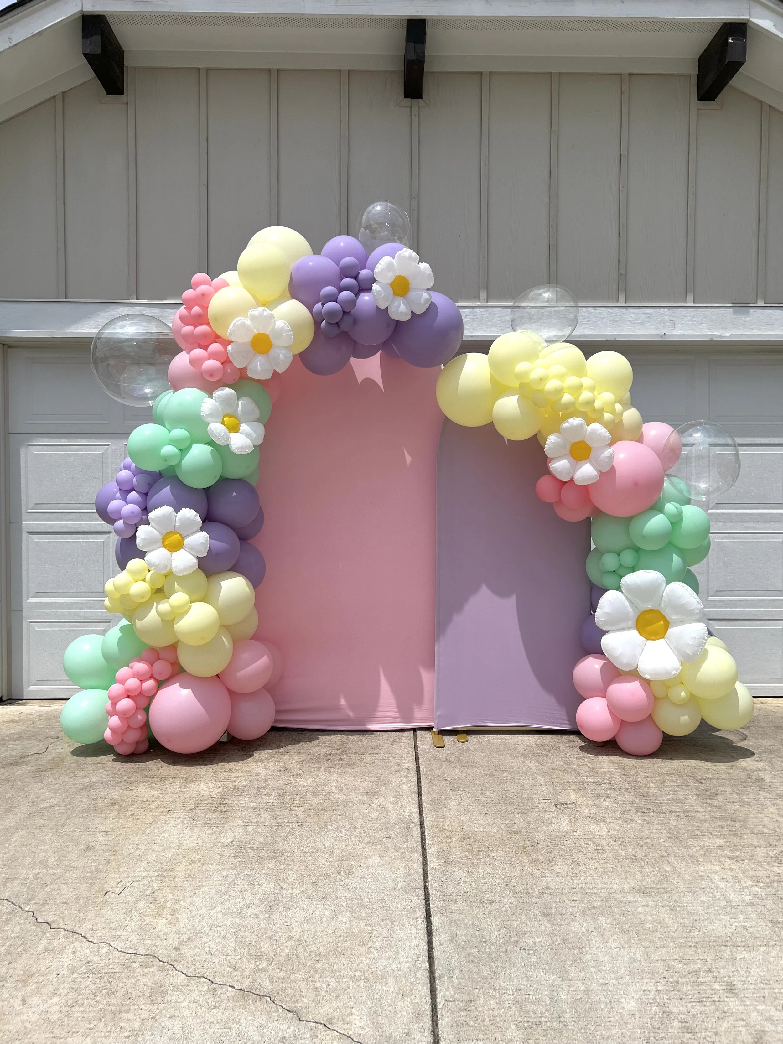 Balloon arch in pastel colors with flowers, transparent globes, pink and purple backdrop, set up outside.