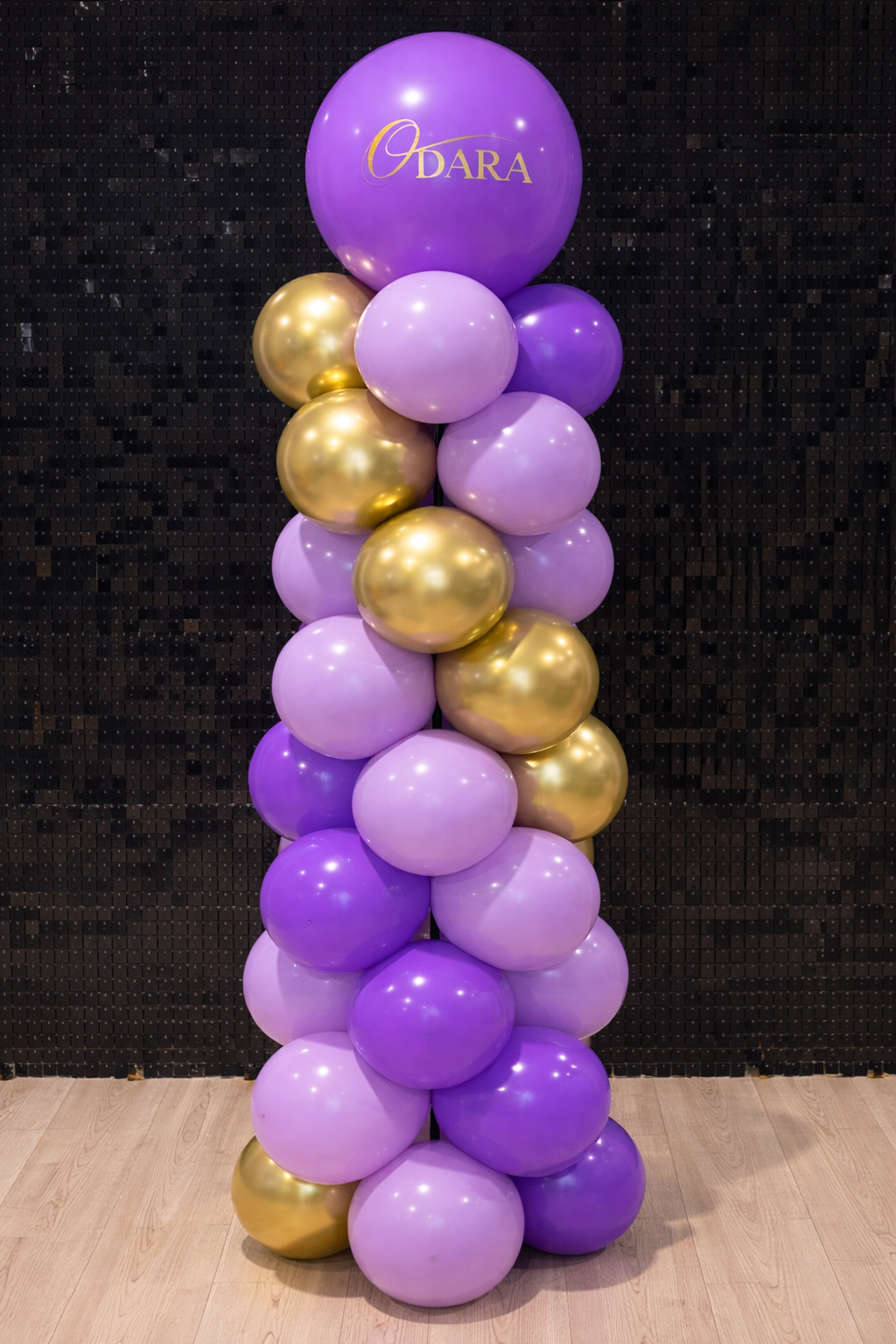 A balloon arrangement consisting of purple, gold, and light purple balloons, with a large purple balloon at the top featuring the word 'Odara' written in gold script.