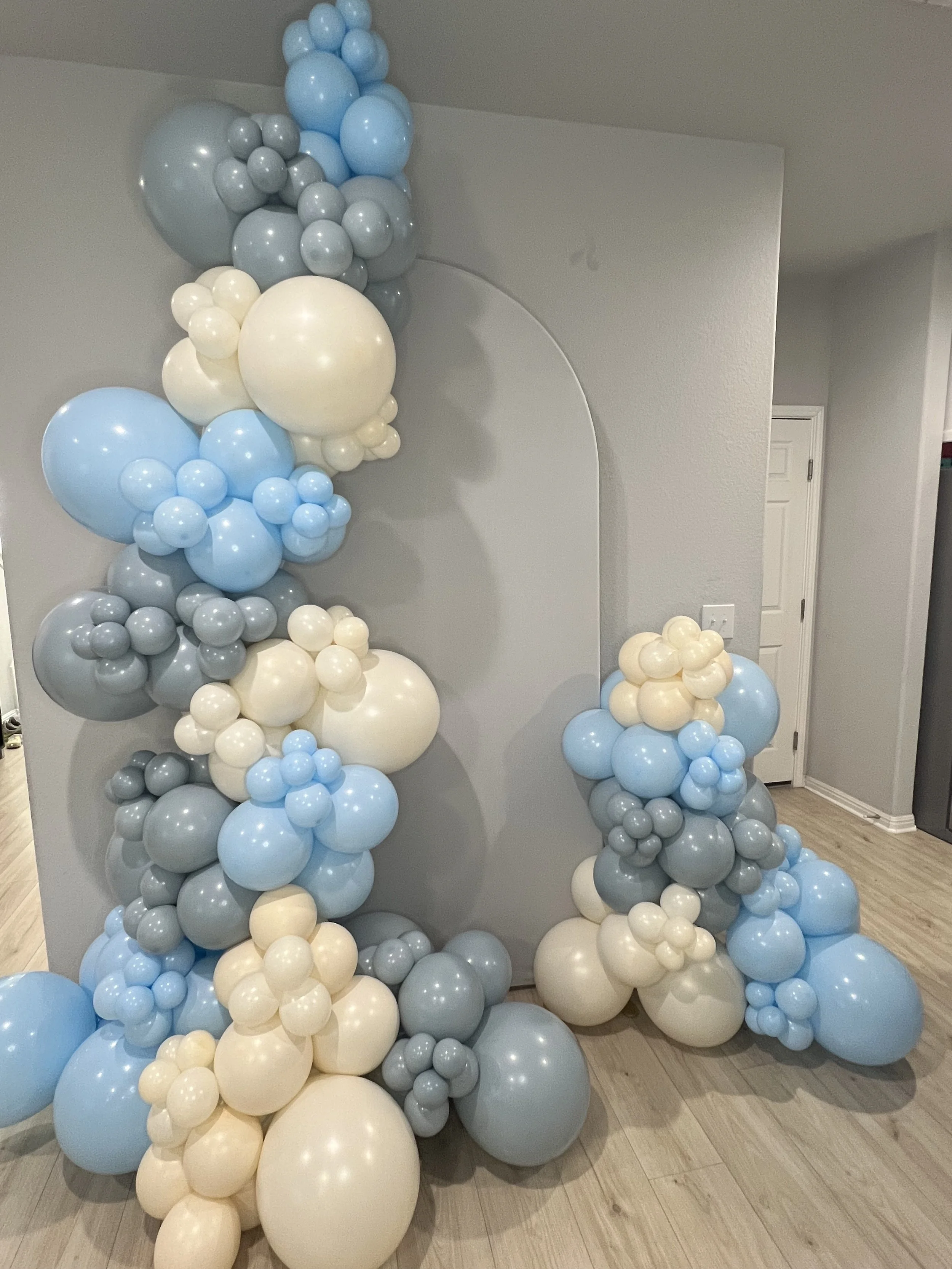Decorative balloon arrangement with clusters of blue, white, and gray balloons in various sizes, set against a plain gray wall.
