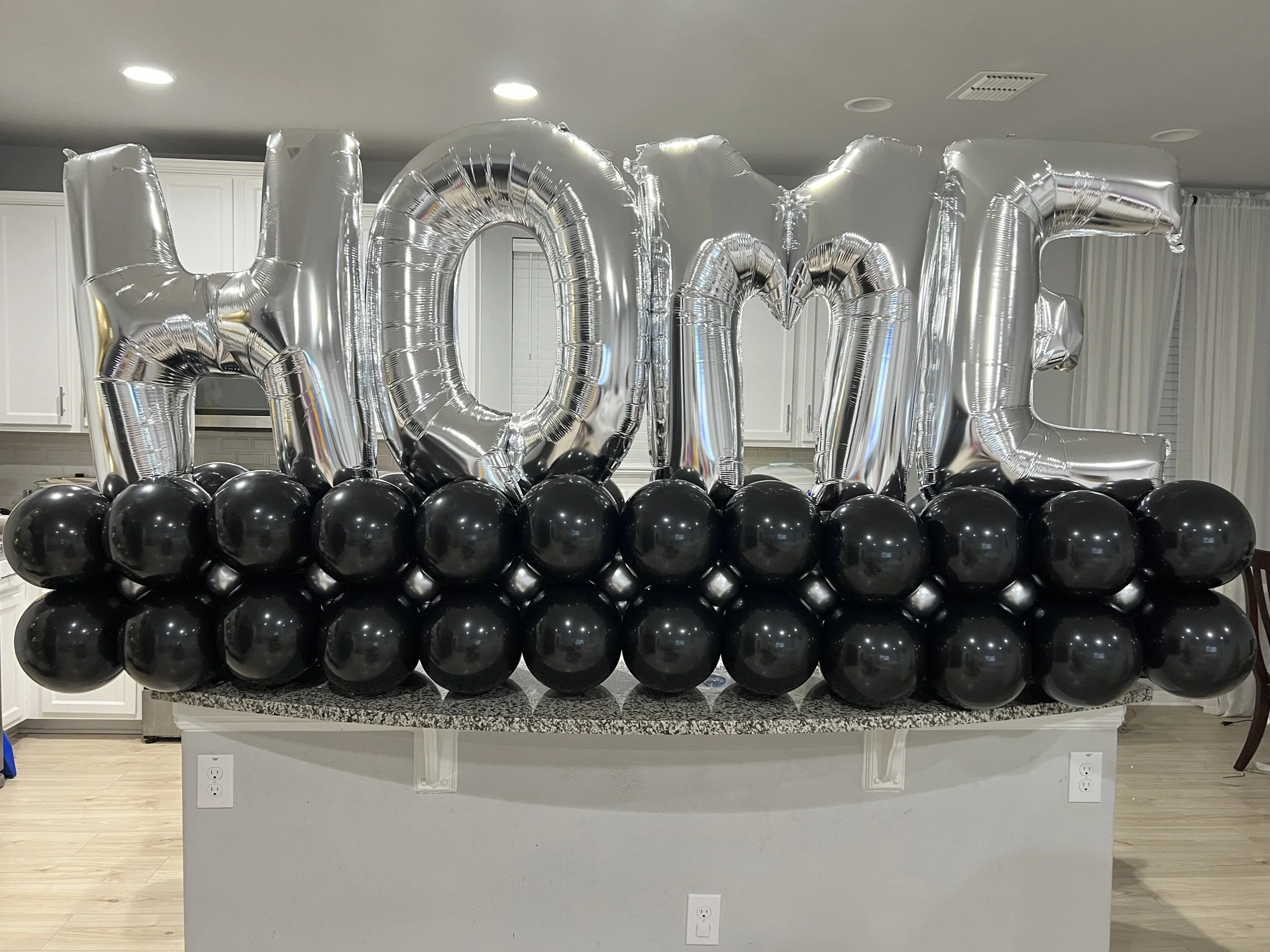 Inflatable foil silver balloons spelling out "HOME" with black balloons underneath on a kitchen island.