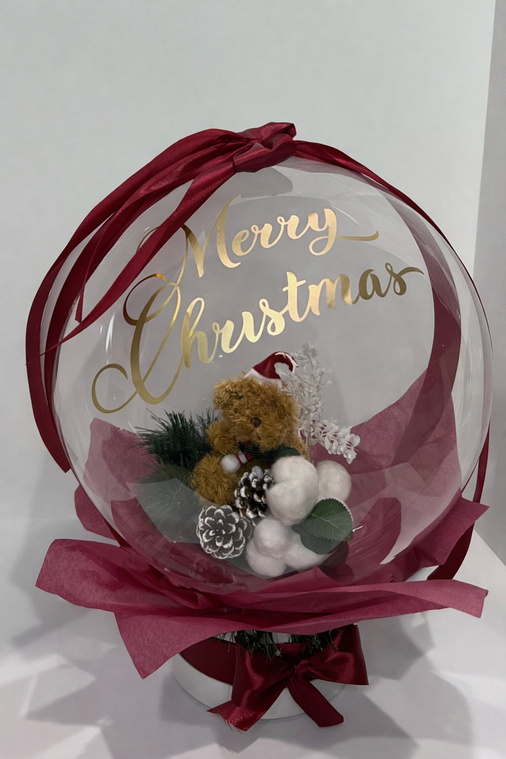 A festive Christmas decoration with a teddy bear wearing a Santa hat inside a bobo balloon, with white cotton balls, pine cones, and green foliage. The balloon has a gold 'Merry Christmas' sign and is wrapped with red ribbons.