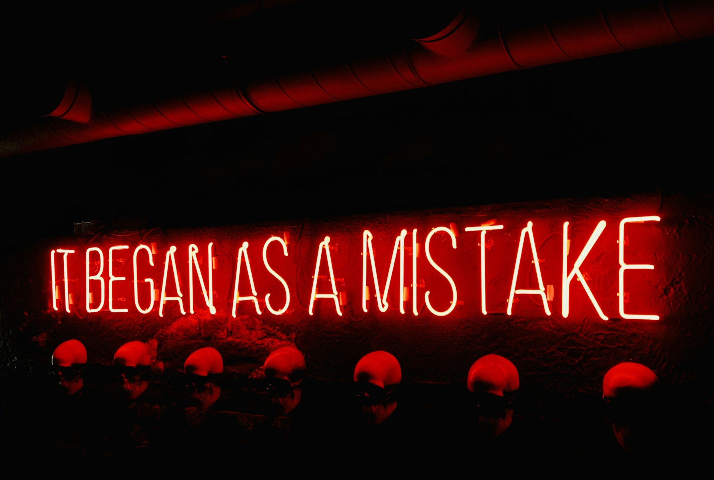 A red neon sign on a dark wall that reads 'IT BEGAN AS A MISTAKE' with a row of indistinct, dark figures lined up underneath it.