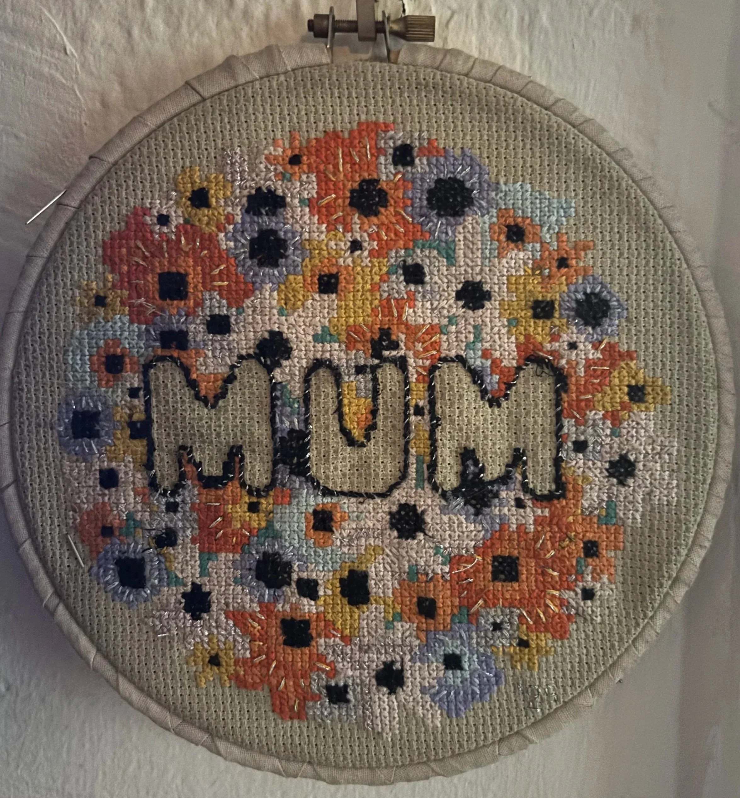 Cross-stitch embroidery of the word 'MOM' surrounded by colorful flowers on beige fabric.