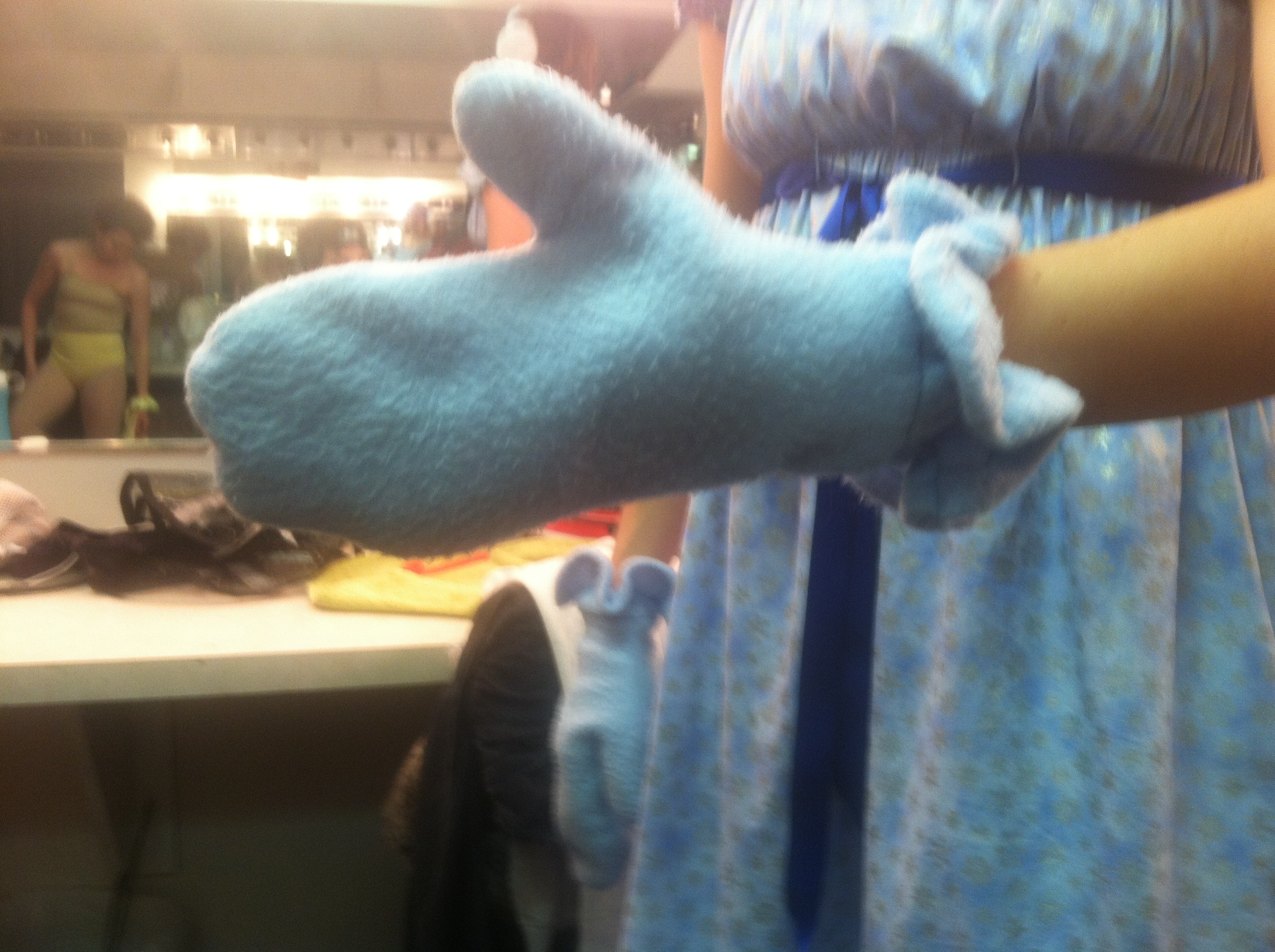 Close-up of a person wearing a blue fleece glove and a blue patterned dress, extending their arm with a gloved hand. In the background, a person in a yellow swimsuit is seen through a mirror in a room that appears to be a kitchen or dining area.