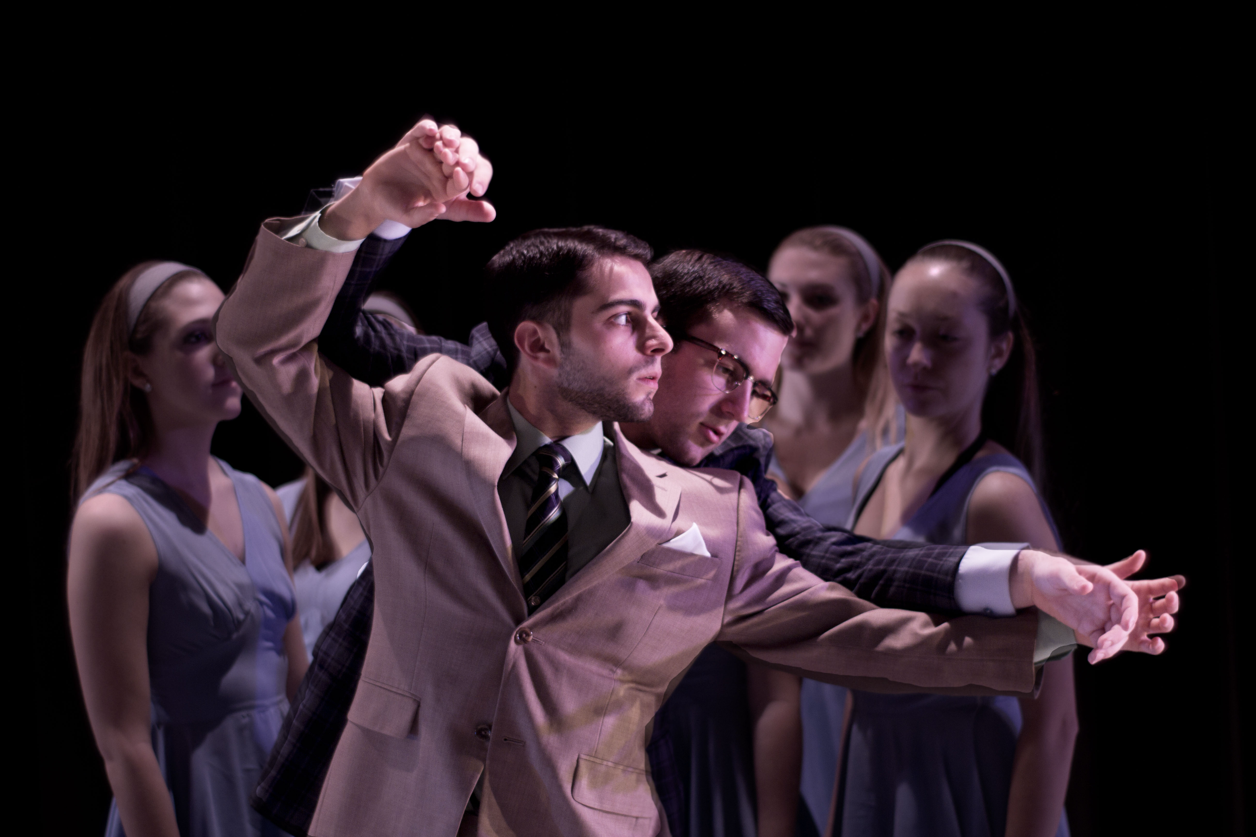 Two men in suits are engaged in a staged fight with multiple young women watching on a dark stage.