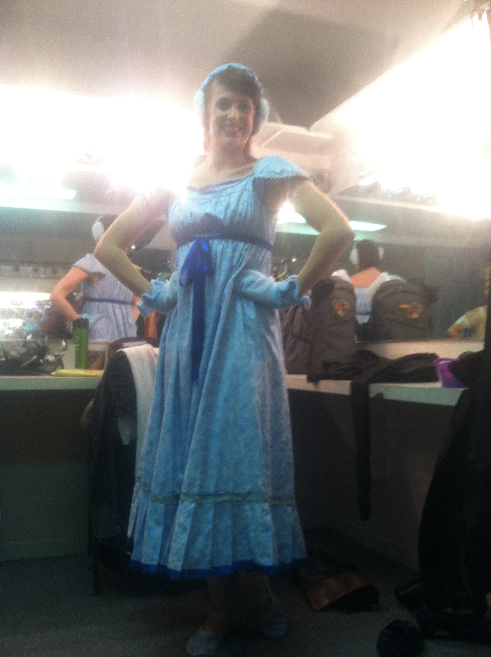 Person dressed in a blue costume with a dress and headpiece, standing in front of a mirror with their hands on their hips, in a dressing room.