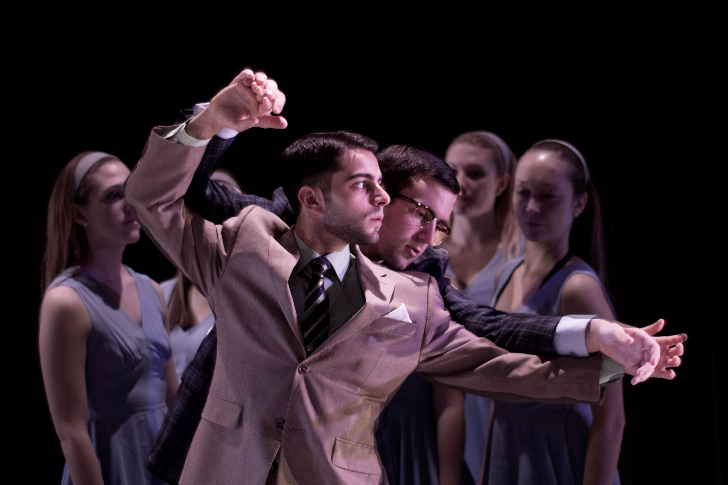 Two men in suits are engaged in a dance with multiple young women watching on a dark stage.
