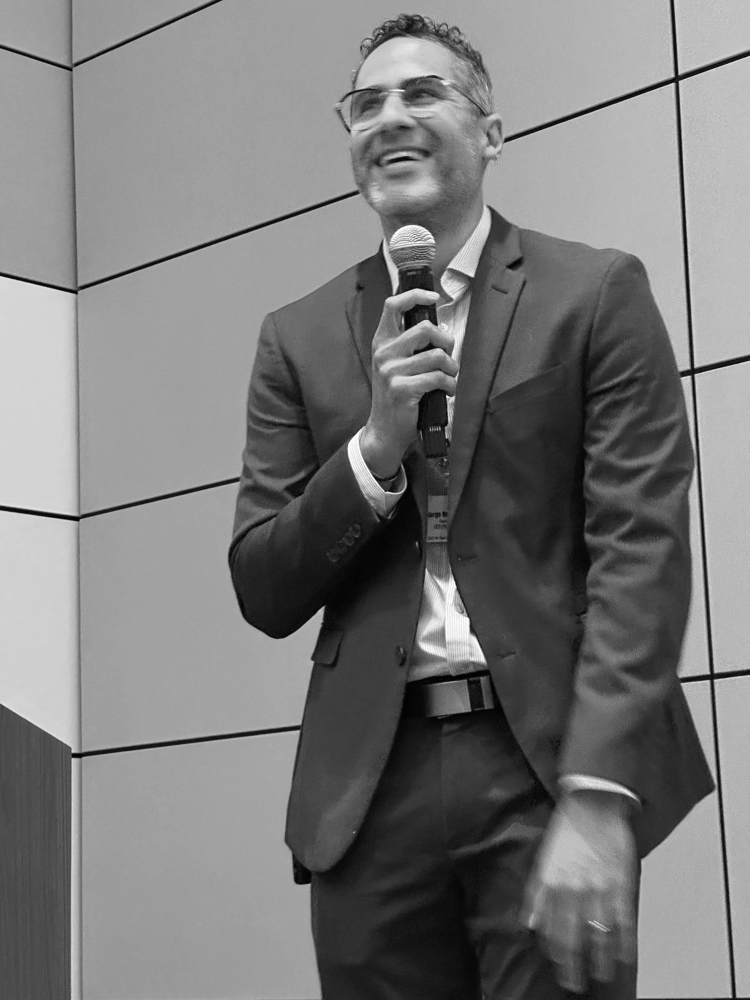 A man in a suit and glasses is smiling and speaking into a microphone at a conference or event.
