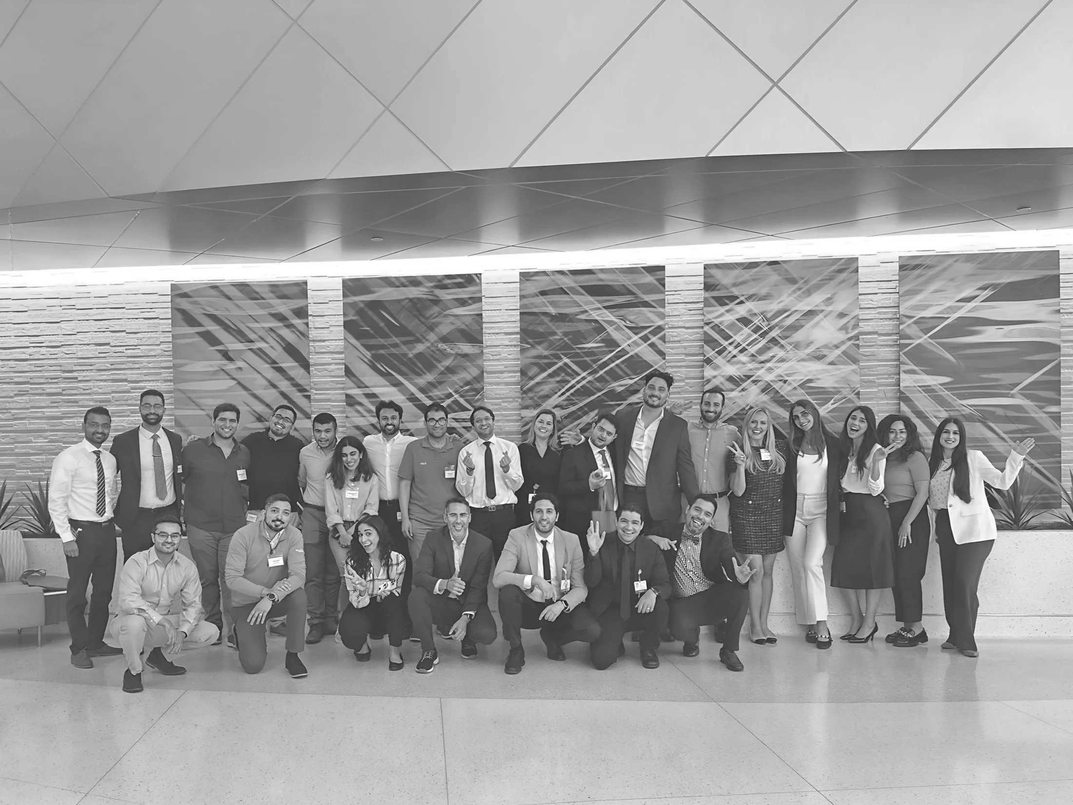 Group of diverse professionals posing together indoors, smiling and making gestures, in front of a decorative wall with abstract patterns.