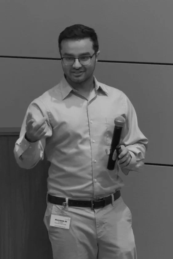 A man wearing glasses, a light-colored button-up shirt, and khakis holding a microphone while gesturing with his left hand, standing in front of a paneled wall at an indoor event.
