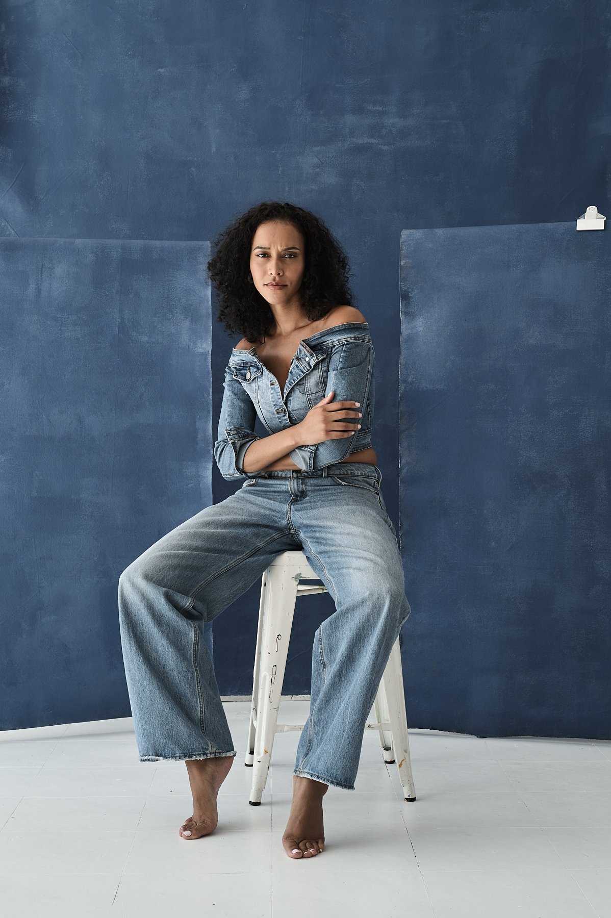 Young woman with curly hair wearing a denim jacket and jeans, sitting barefoot on a stool against a blue wall.