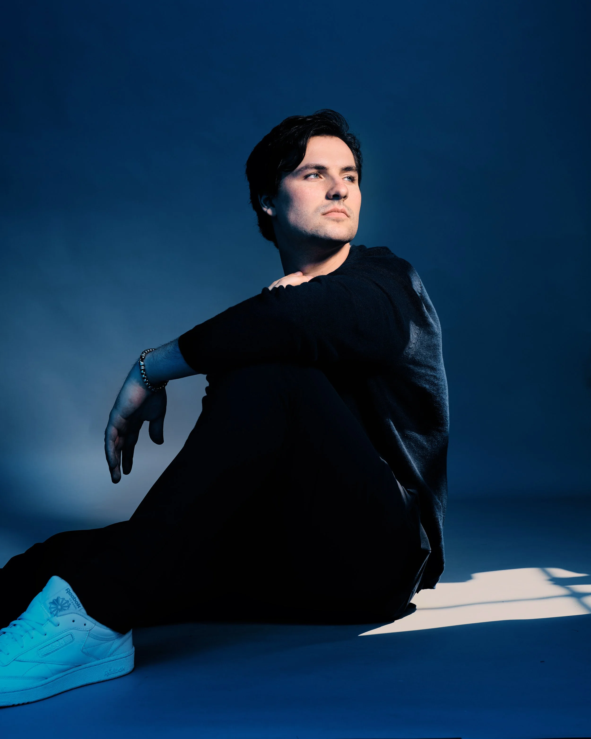 Young man sitting on the floor against a blue background, wearing a black sweater, black pants, and white sneakers, looking off to the side.