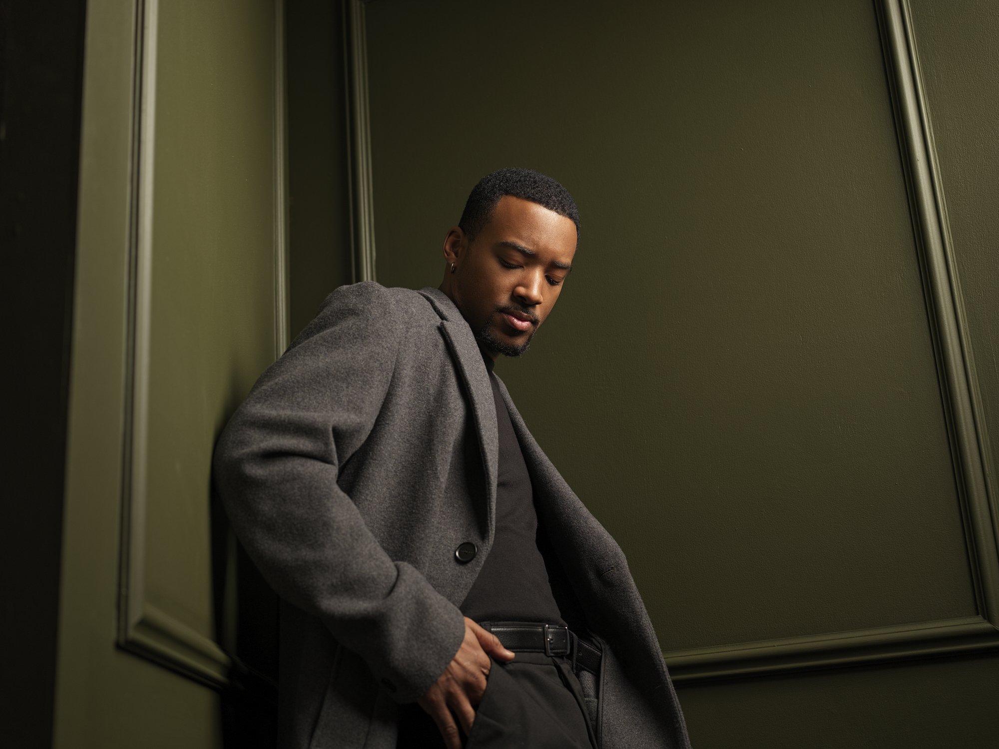 A man leaning against a green wall, wearing a gray coat and black outfit with his hand in his pocket, looking down.