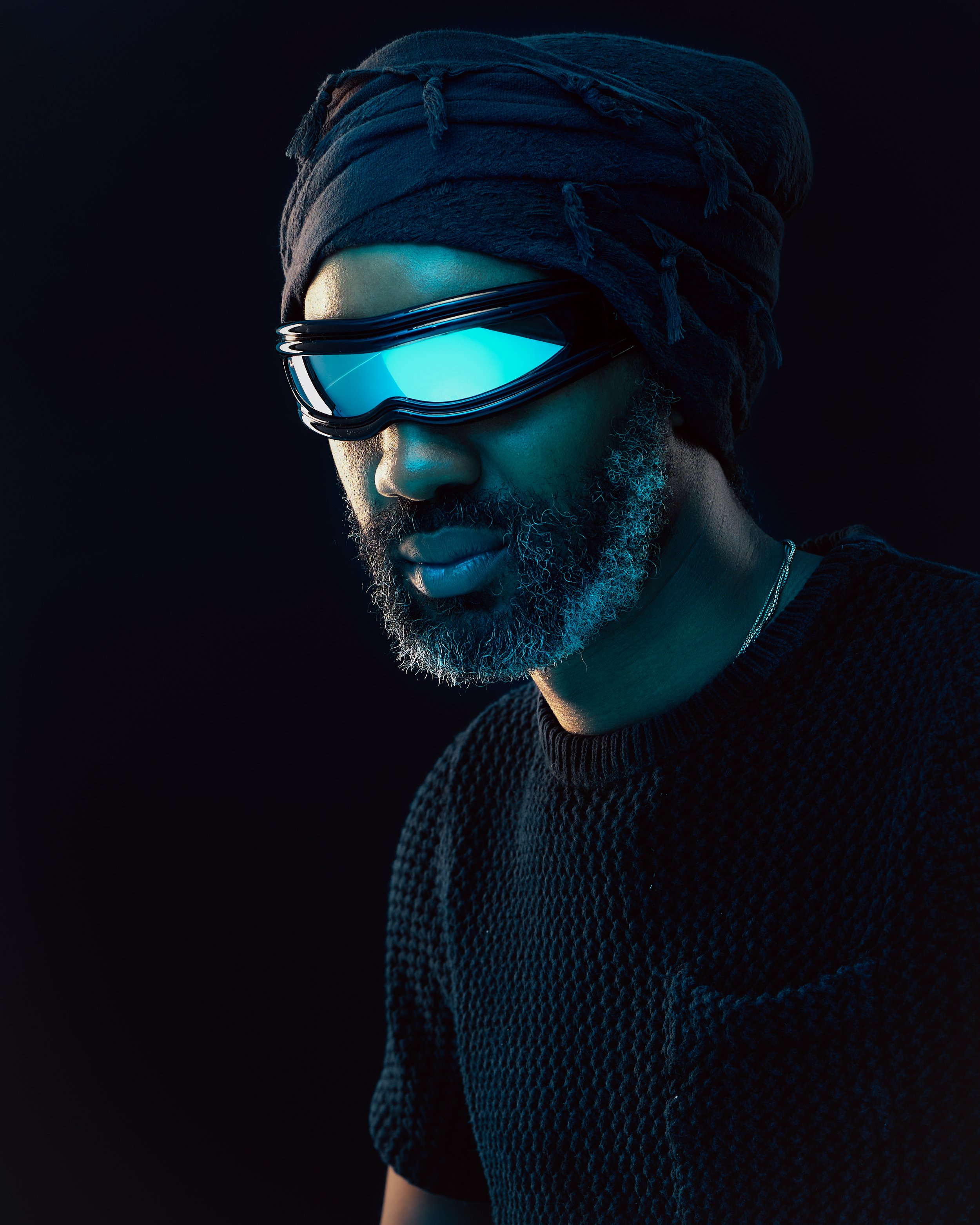 A man wearing a black turban and reflective futuristic goggles, with a beard, dark clothing, and a dark background.