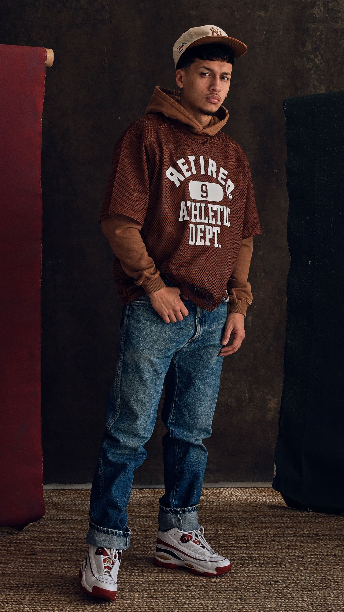 A young man wearing a beige baseball cap, a brown athletic hoodie layered over a tan long sleeve shirt, blue jeans, and white sneakers with red and black accents, standing against a dark background.