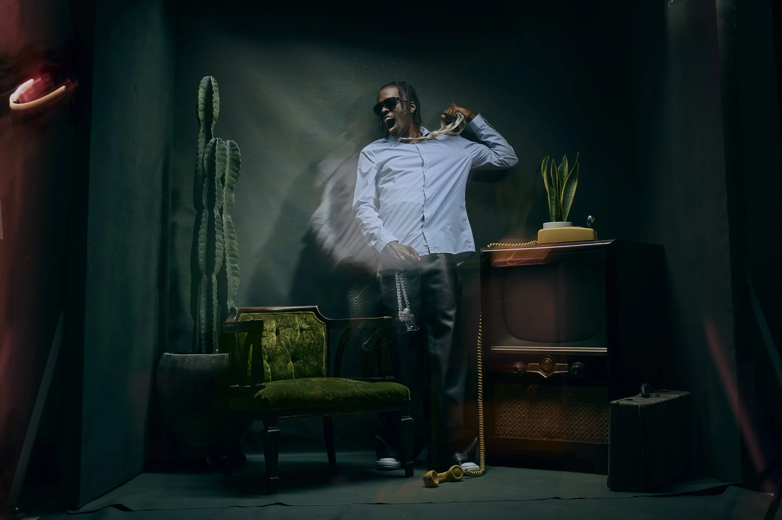 Person with sunglasses, dressed in a white shirt and leather pants, in a dimly lit room with vintage furniture, a cactus, and a television, striking an expressive pose.