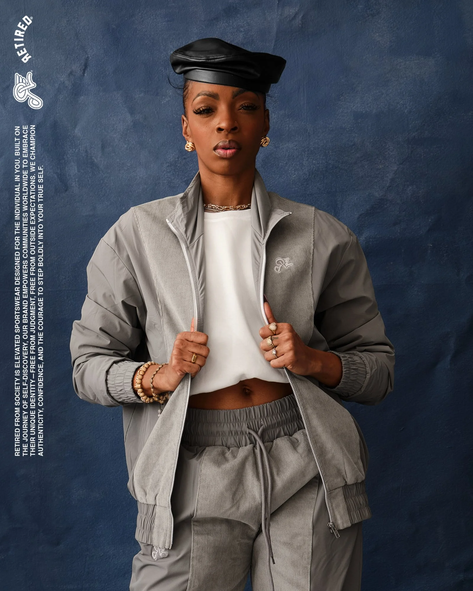 A woman dressed in a sporty gray tracksuit and white crop top stands against a textured dark blue background.