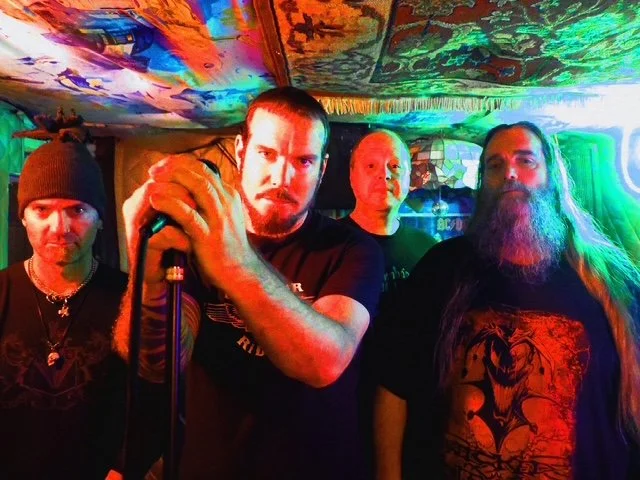 Four men standing in a dimly lit room with colorful, psychedelic lighting and artwork on the ceiling, one holding a microphone, suggesting a musical performance or band.