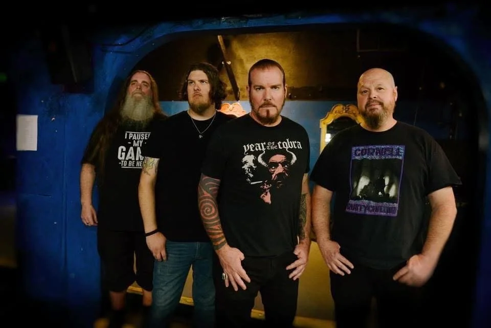 Four men standing together in a dimly lit room with blue and yellow wall lighting, wearing dark band T-shirts.