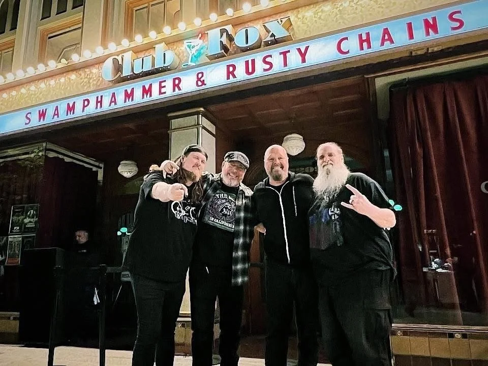 Four people standing in front of a stage with a sign that reads 'Club Fox' and 'Swamp Hammer & Rusty Chains'. They are posing for the photo, with one person making a peace sign. The setting appears to be a bar or music venue.