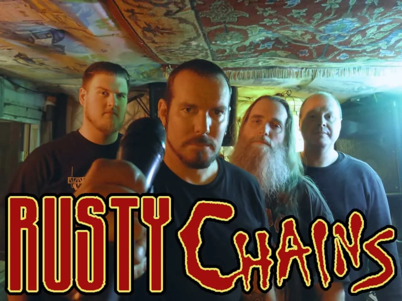 Four men standing in a room with a decorative ceiling, one of whom is holding a microphone. The text overlay reads "Rusty Chains" in bold yellow and red letters.
