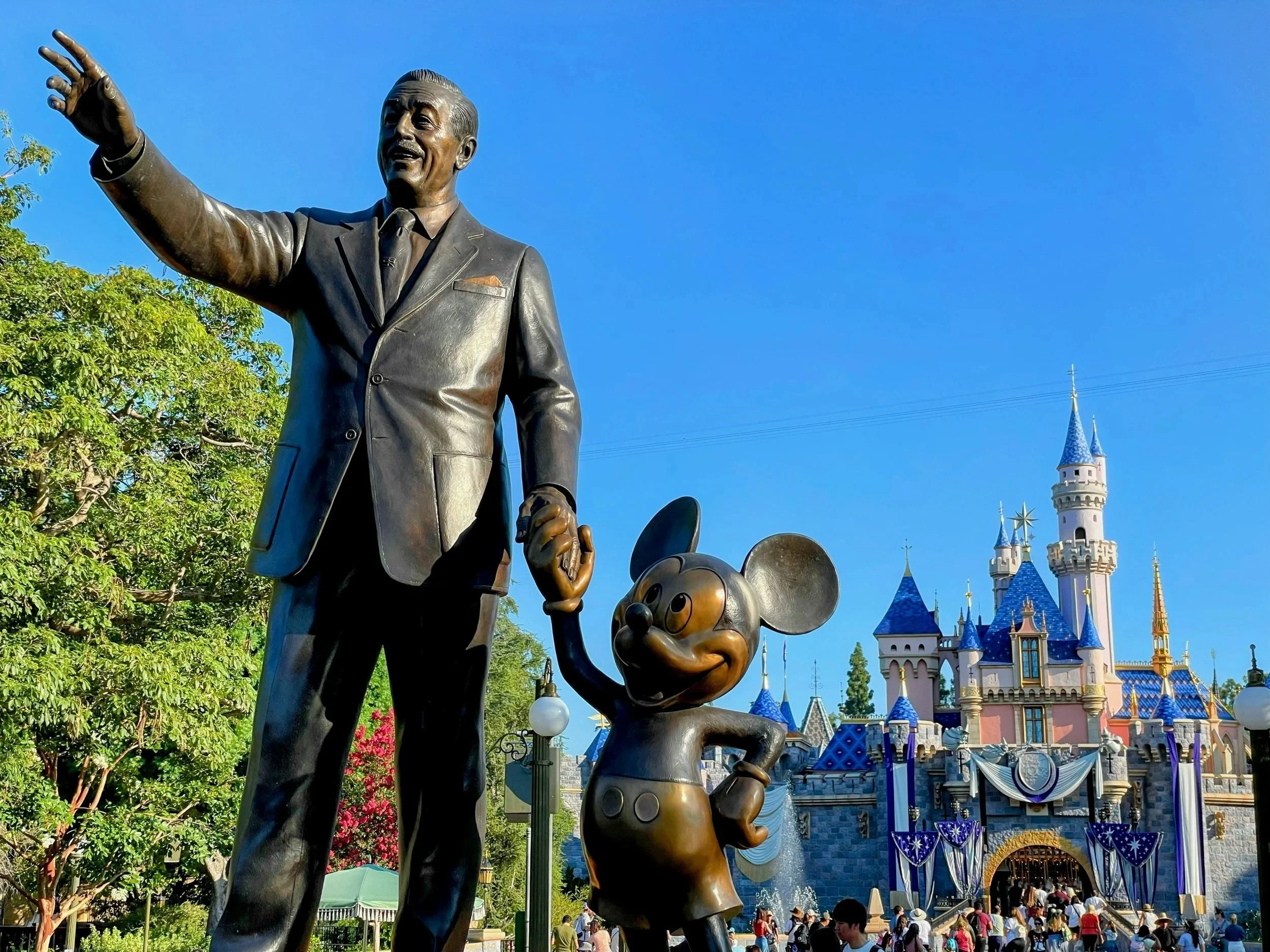 Statue of Walt Disney with Mickey Mouse at Disneyland, with a castle in the background, under a clear blue sky.