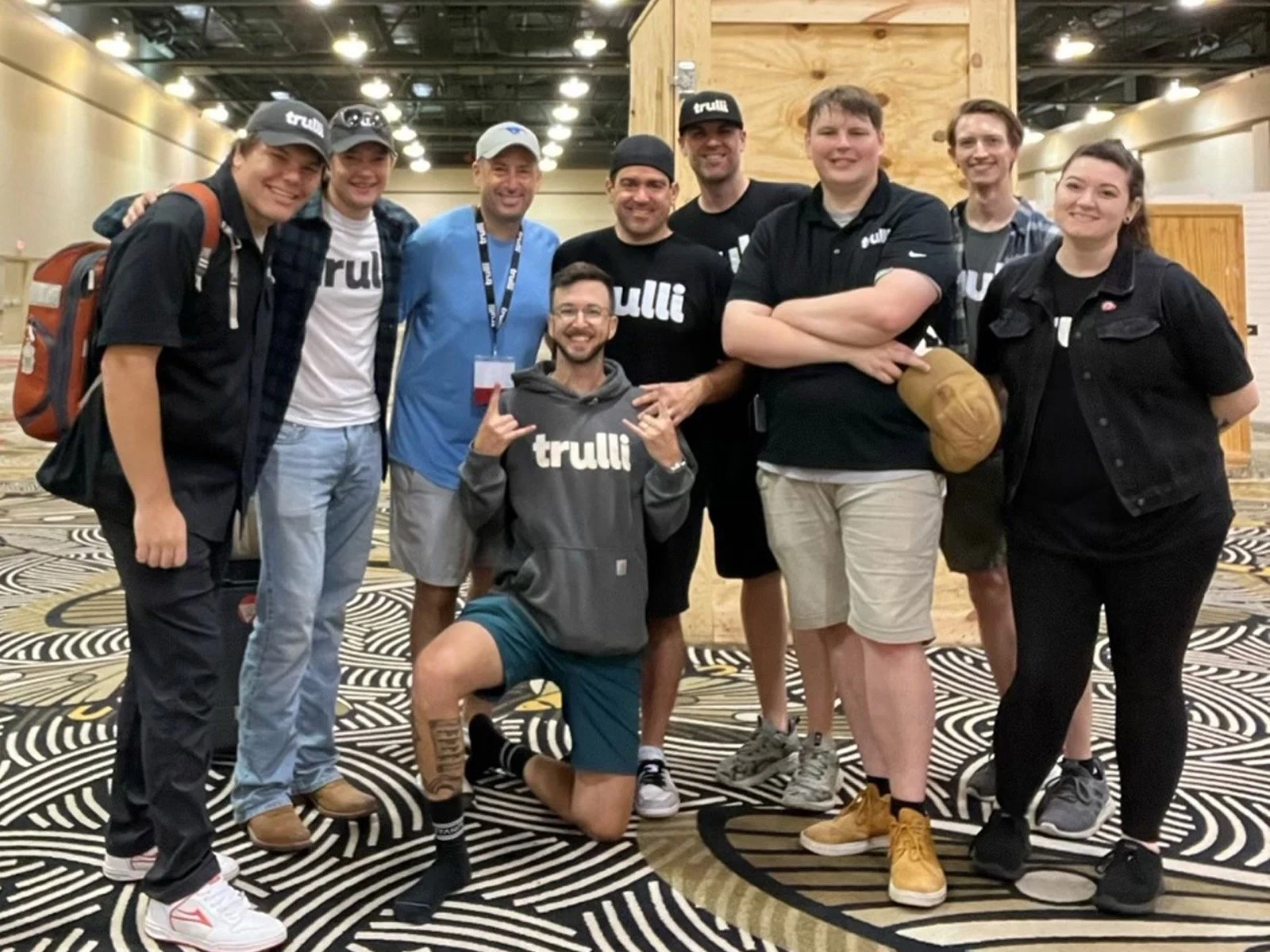 Group of nine young adults posing together indoors, some wearing black tulli shirts, smiling and making gestures, with patterned carpet and wooden walls in the background.