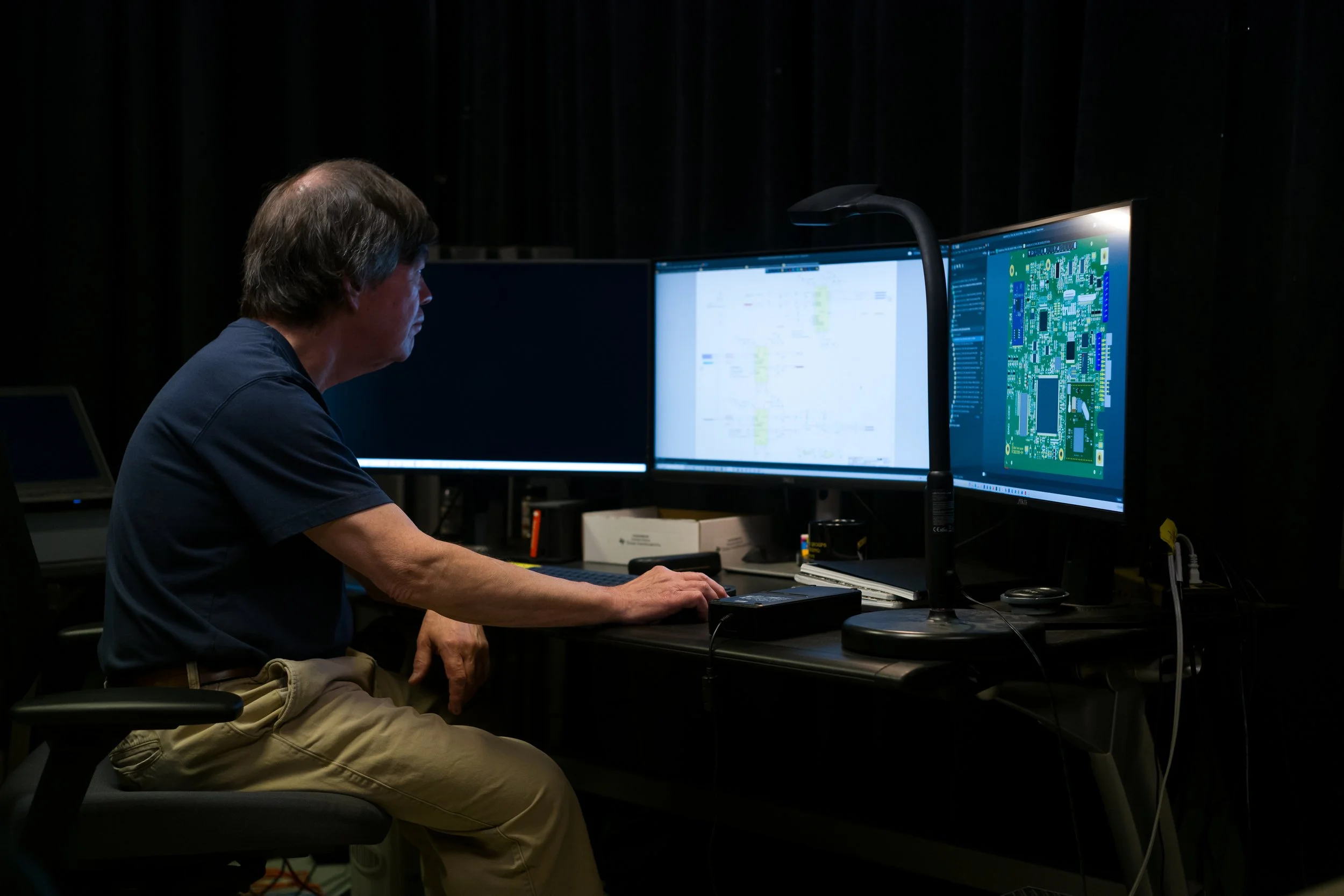 Man in a dark room working at a desk with three computer monitors displaying technical drawings and circuit diagrams.
