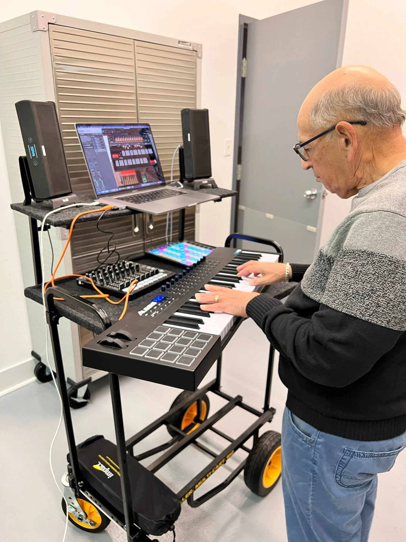 An elderly man playing a digital keyboard with music production equipment, a laptop with music software, and speakers on a music stand.