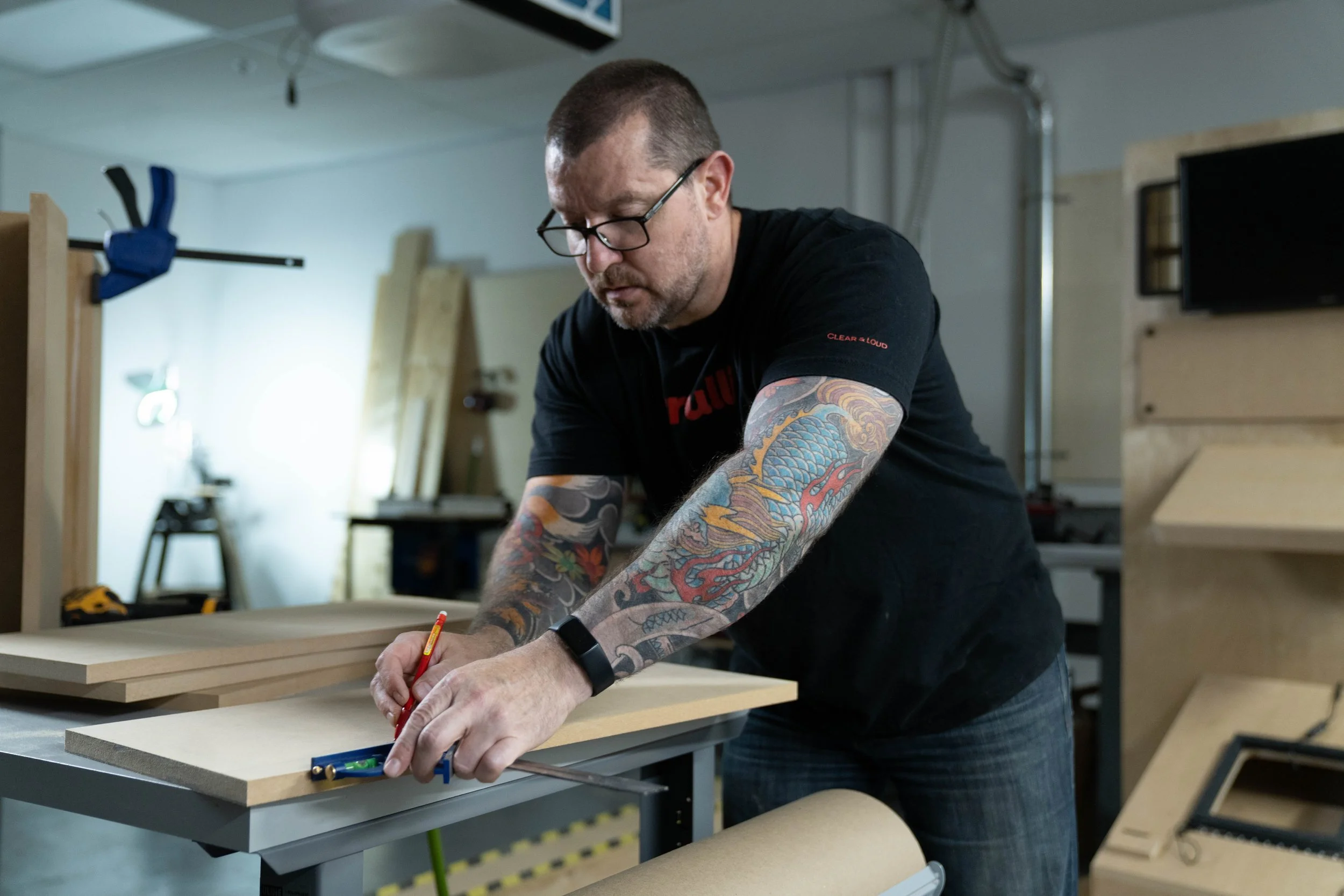 A man with tattoos measuring and marking a piece of wood in a woodworking shop.
