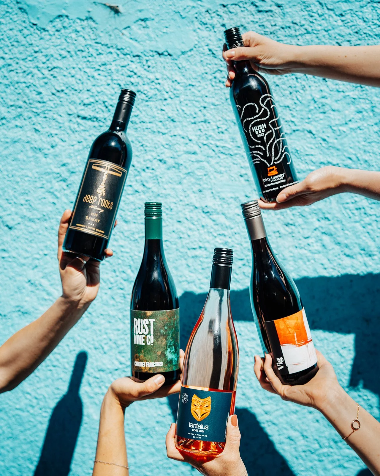Multiple hands holding bottles of wine and alcohol against a textured blue wall, with some bottles stacked and others being lifted.