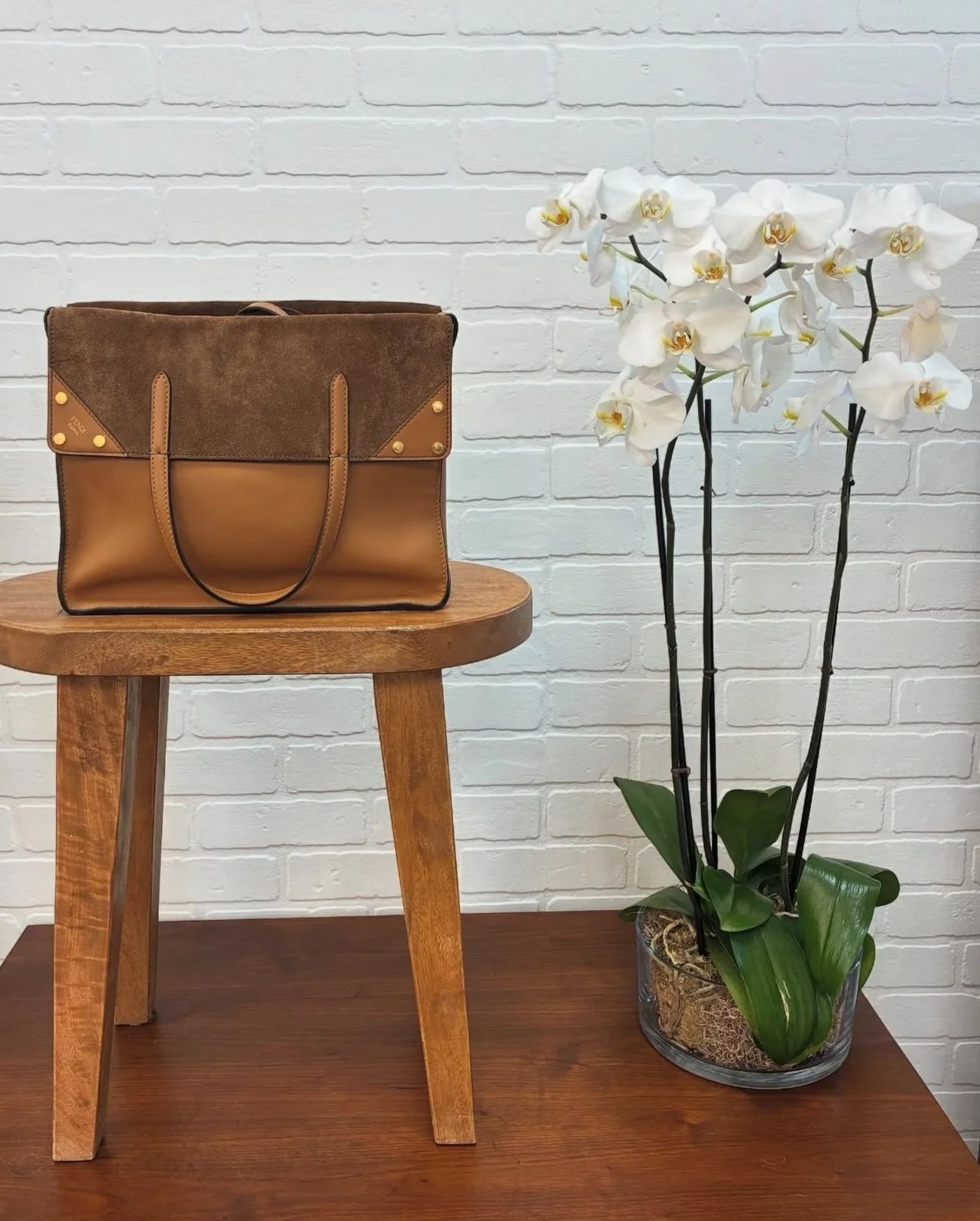 A brown leather handbag with a suede flap is placed on a wooden stool, next to an air-potted white orchid plant with green leaves, against a white brick wall.