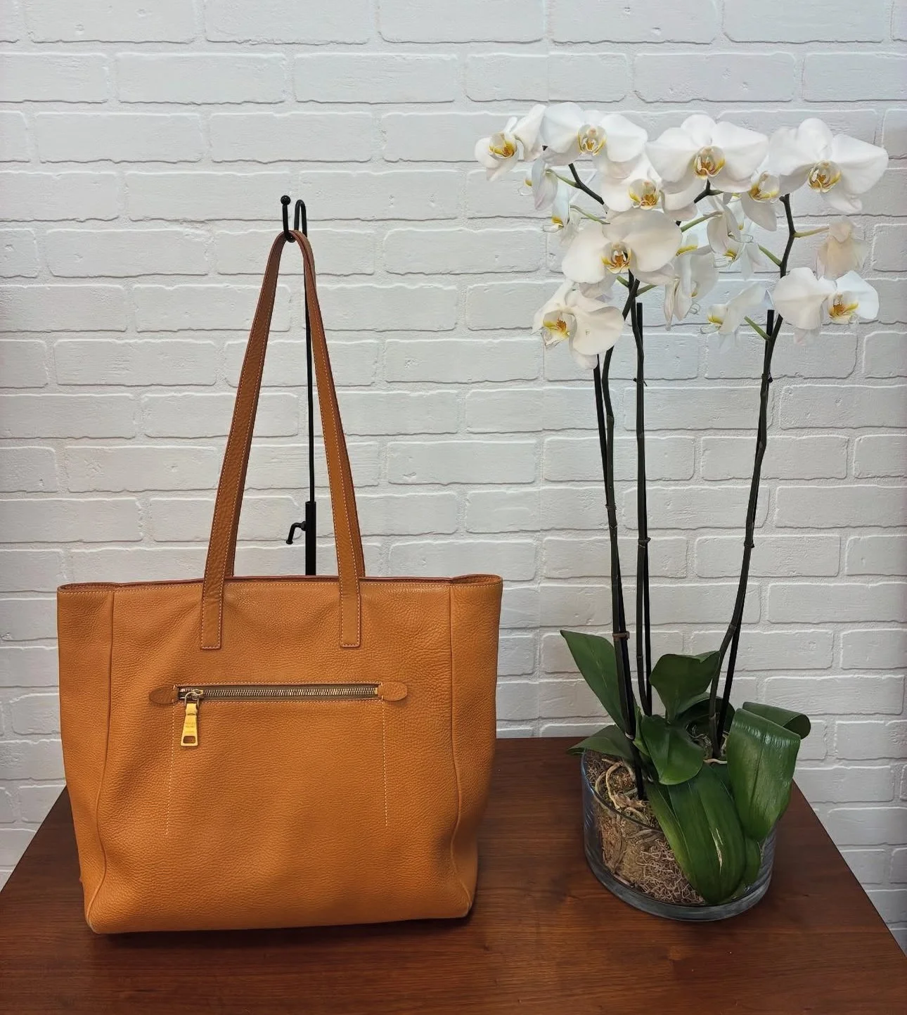 A large tan leather tote bag with a zipper pocket sits on a wooden surface next to a potted orchid with white flowers and green leaves against a white brick wall background.