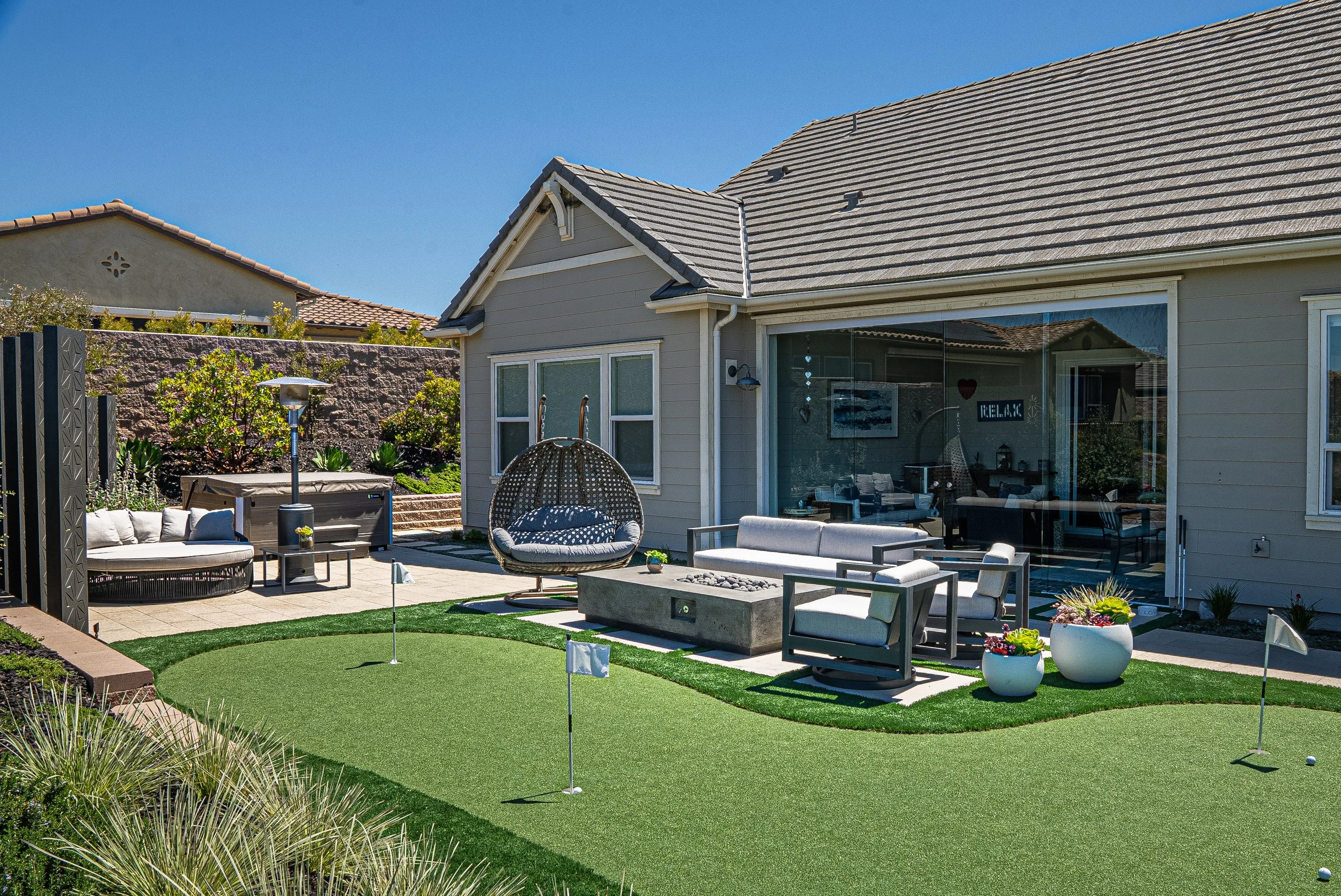 Backyard with putting green, outdoor furniture, and a hot tub, adjacent to a house with a sliding glass door and windows, under a clear blue sky.